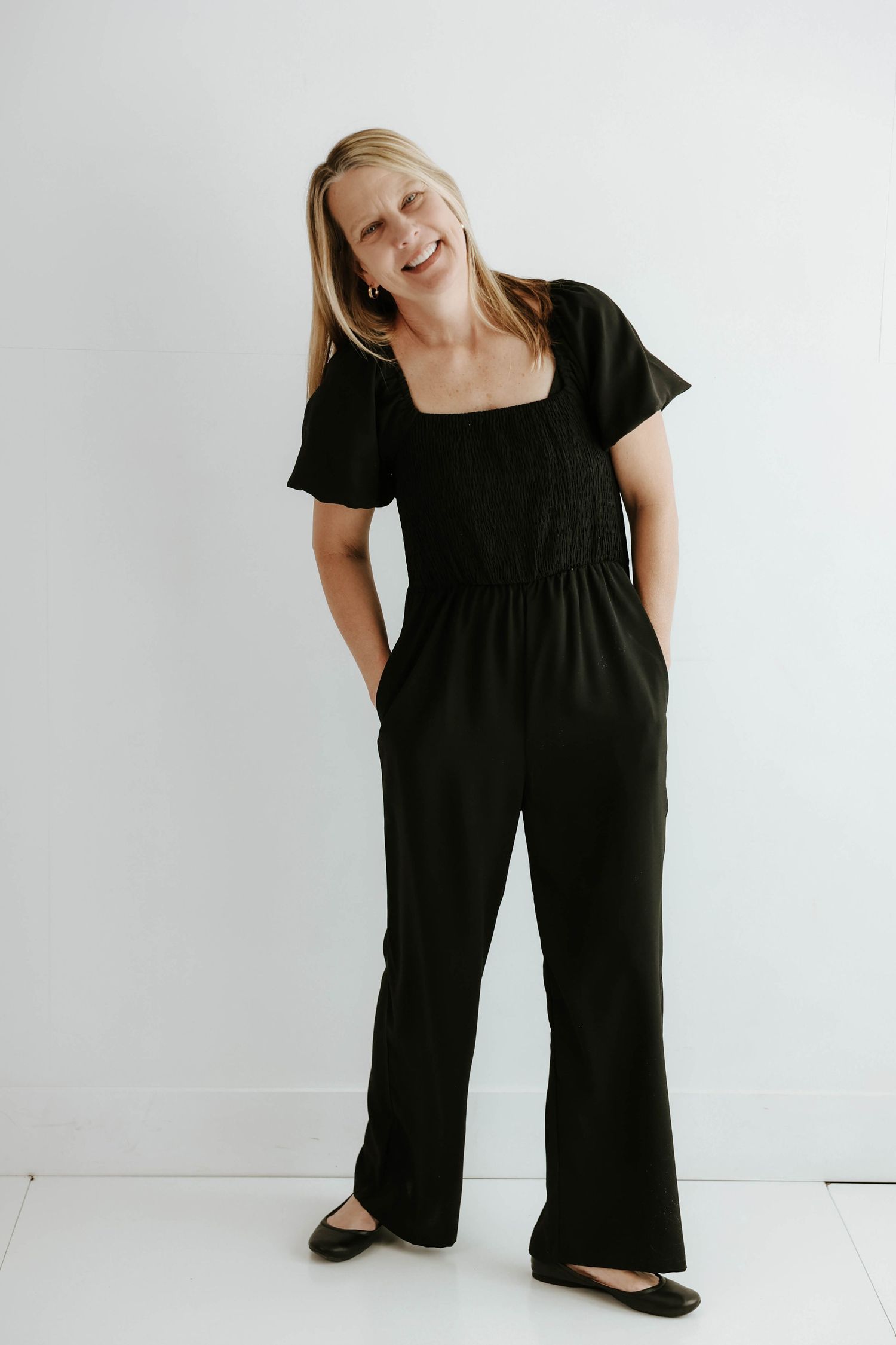 A person wearing a casual black jumpsuit and flats poses cheerfully against a white wall.