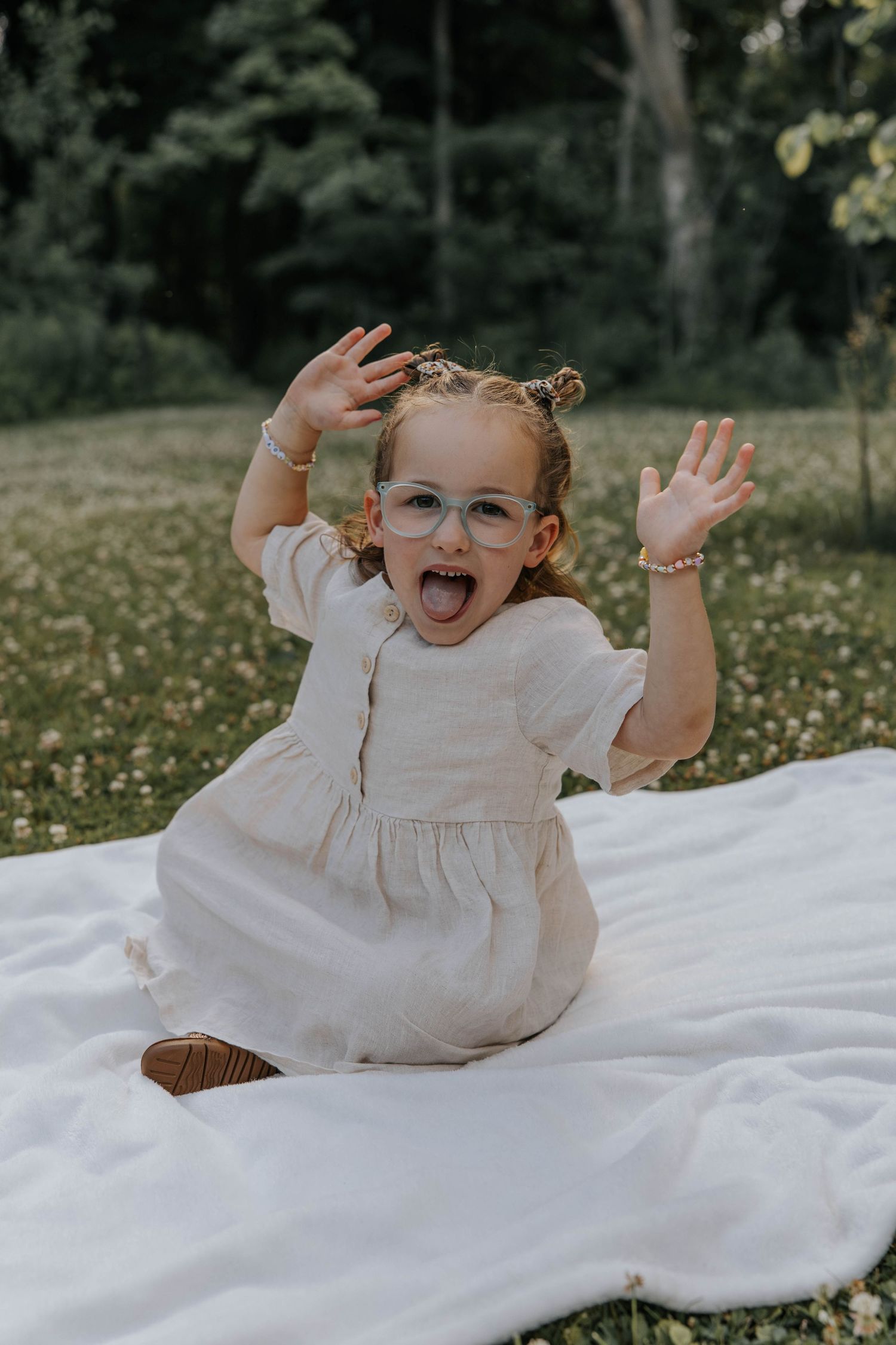 A joyful child in a white dress lies on a white blanket outdoors surrounded by scattered wildflowers.