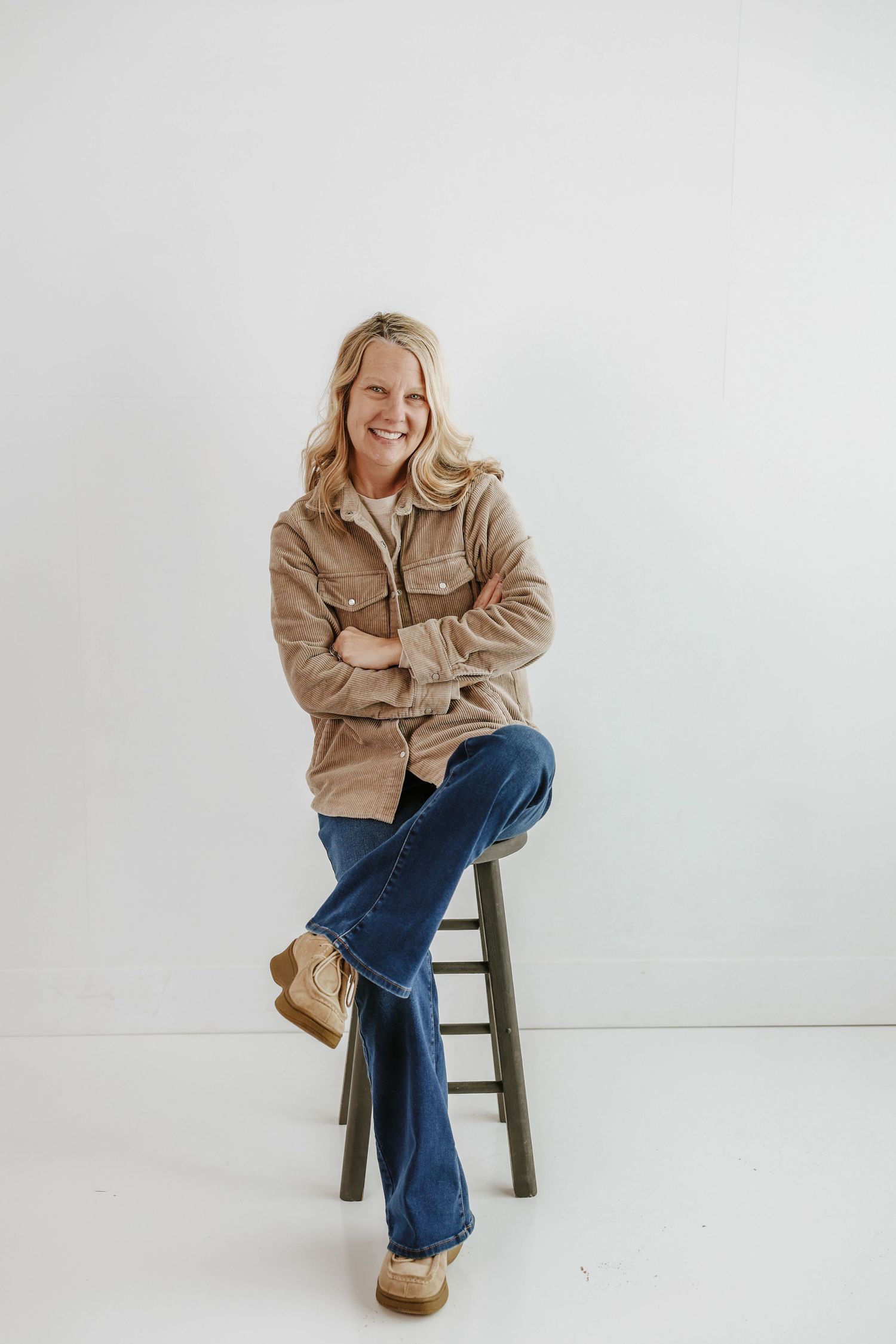 Model in cozy beige jacket and flared jeans poses on wooden stool against white backdrop.