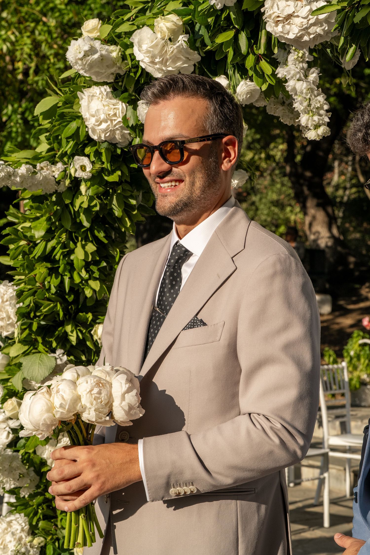 A person in a light gray suit and sunglasses smiles while holding flowers at an outdoor event.