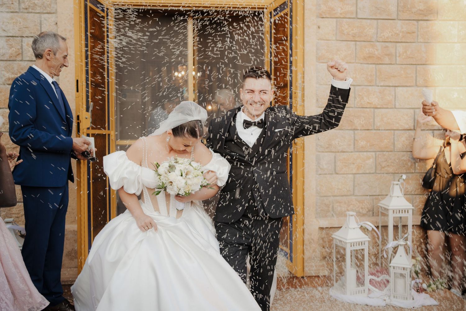 Newlyweds walk through a shower of white rice being thrown by wedding guests outside a stone church.