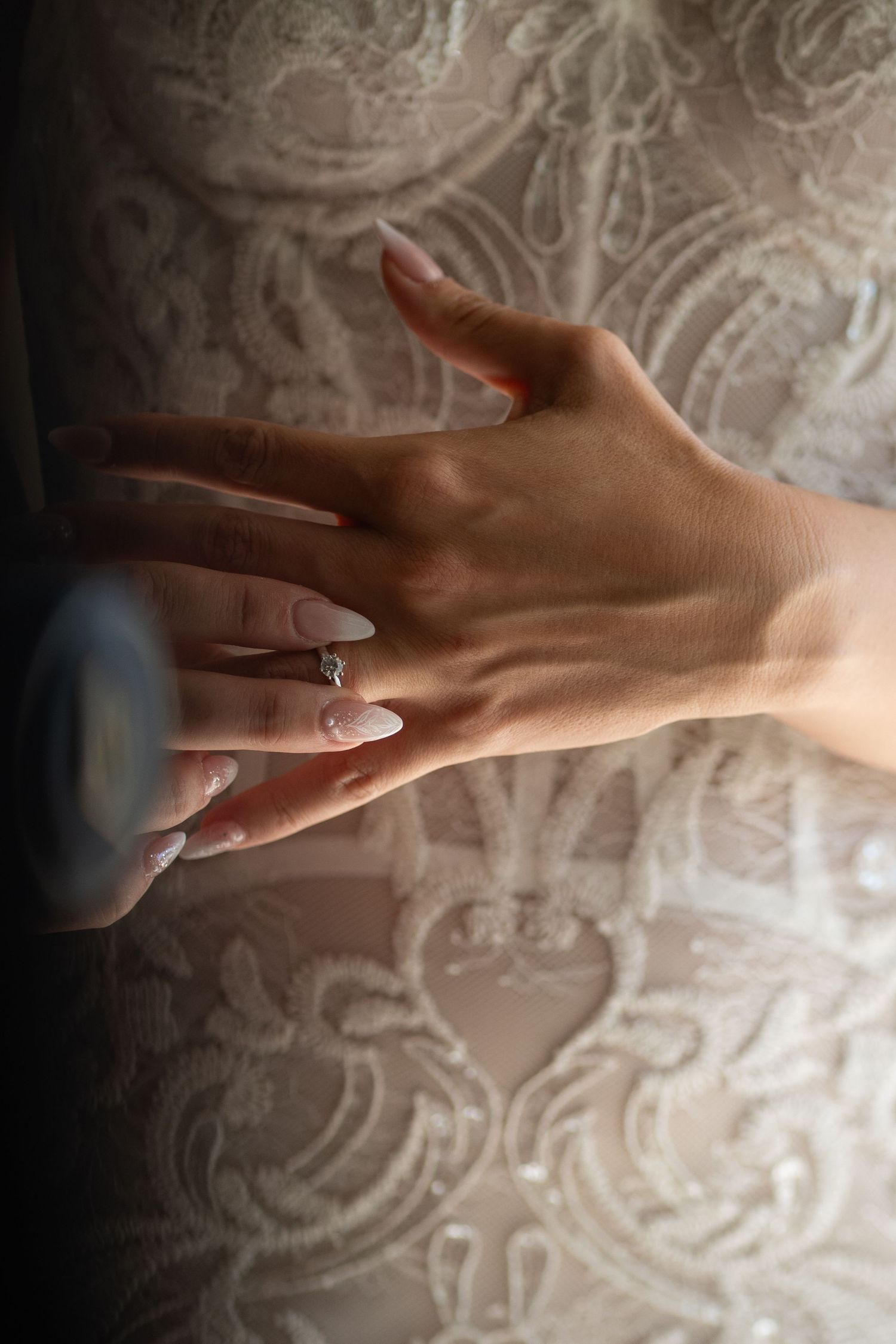 Close-up of manicured hand with engagement ring against lace pattern background.