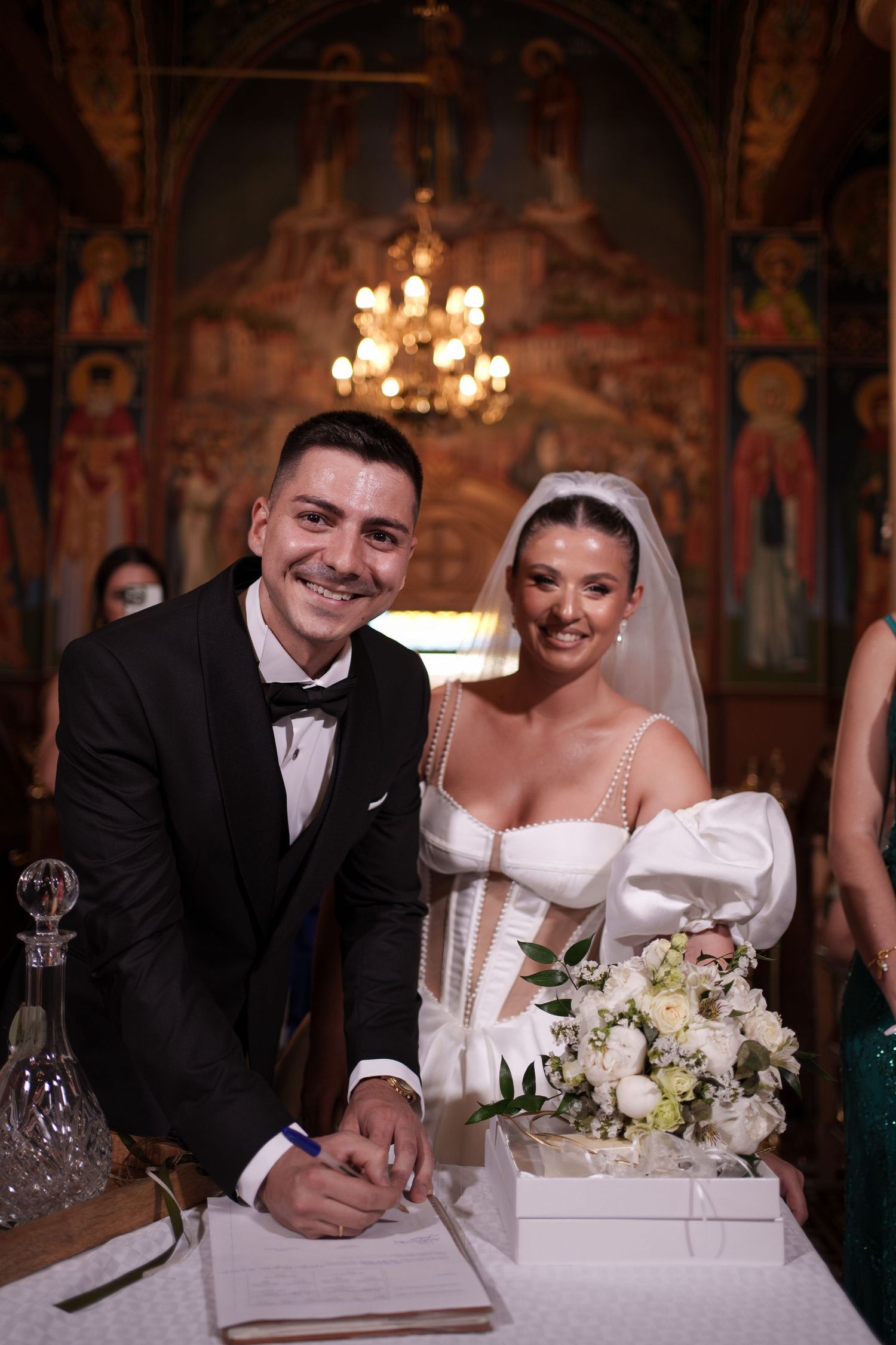 A couple signing their marriage documents during a wedding ceremony in an ornate church setting with chandeliers.