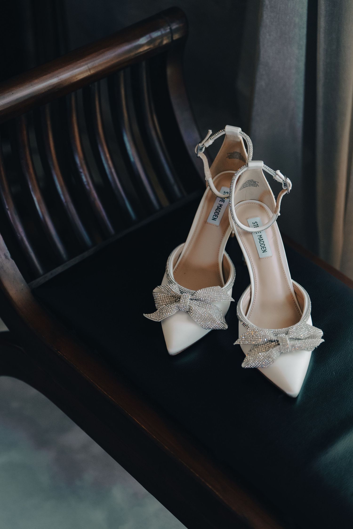 White bridal heels with sparkly bow details rest on a dark wooden chair.
