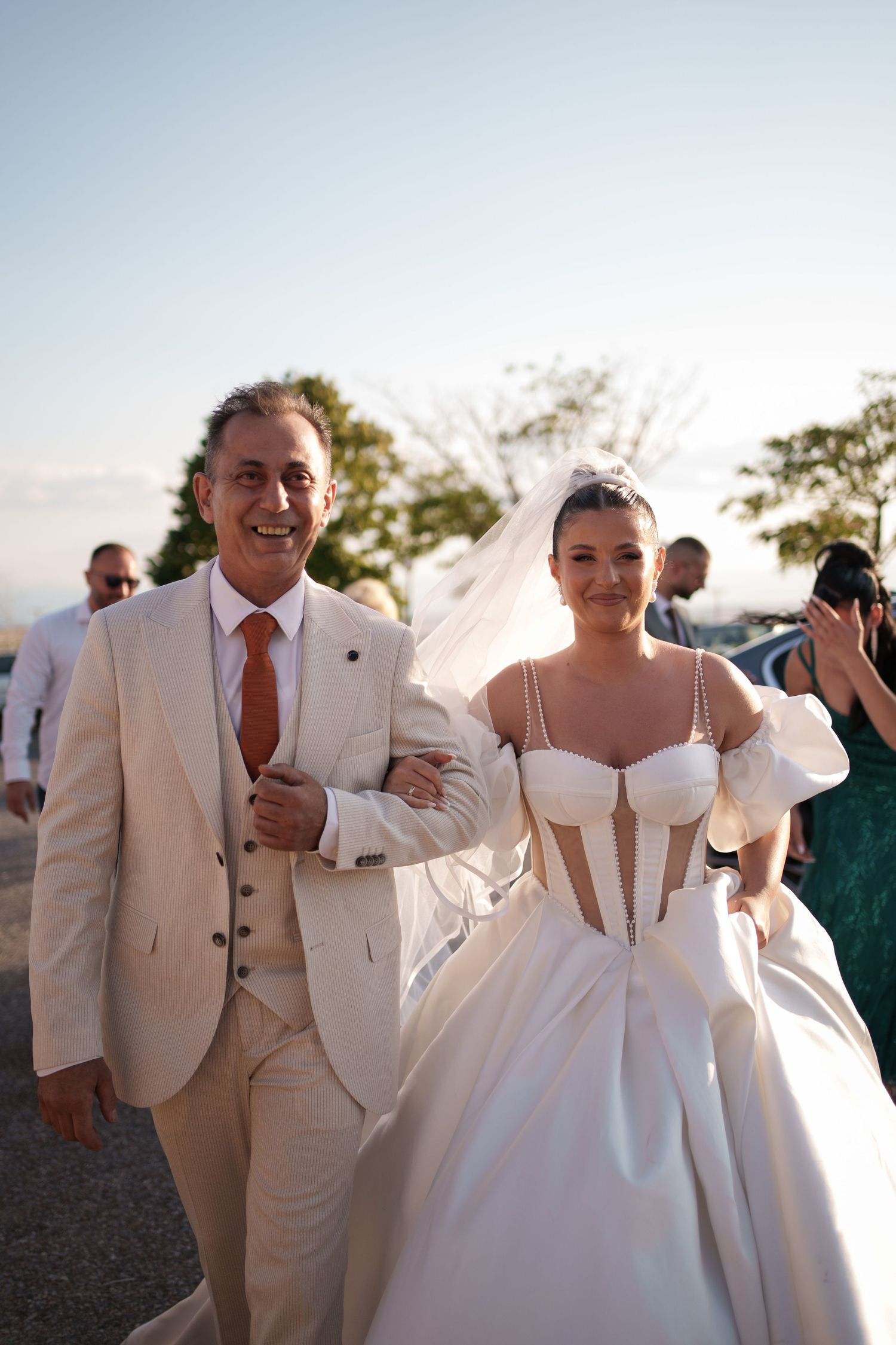 Wedding party walks and laughs together outdoors in beige suits and white dress during sunset celebration.