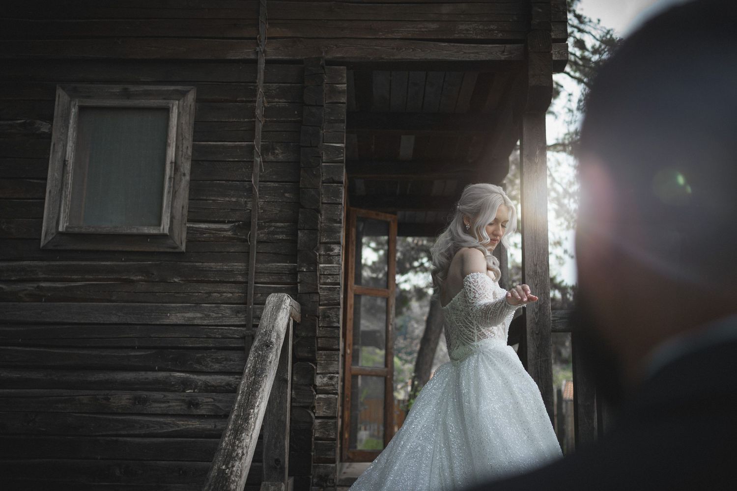 A bride in a white gown stands on the porch of a rustic wooden cabin on her wedding day.