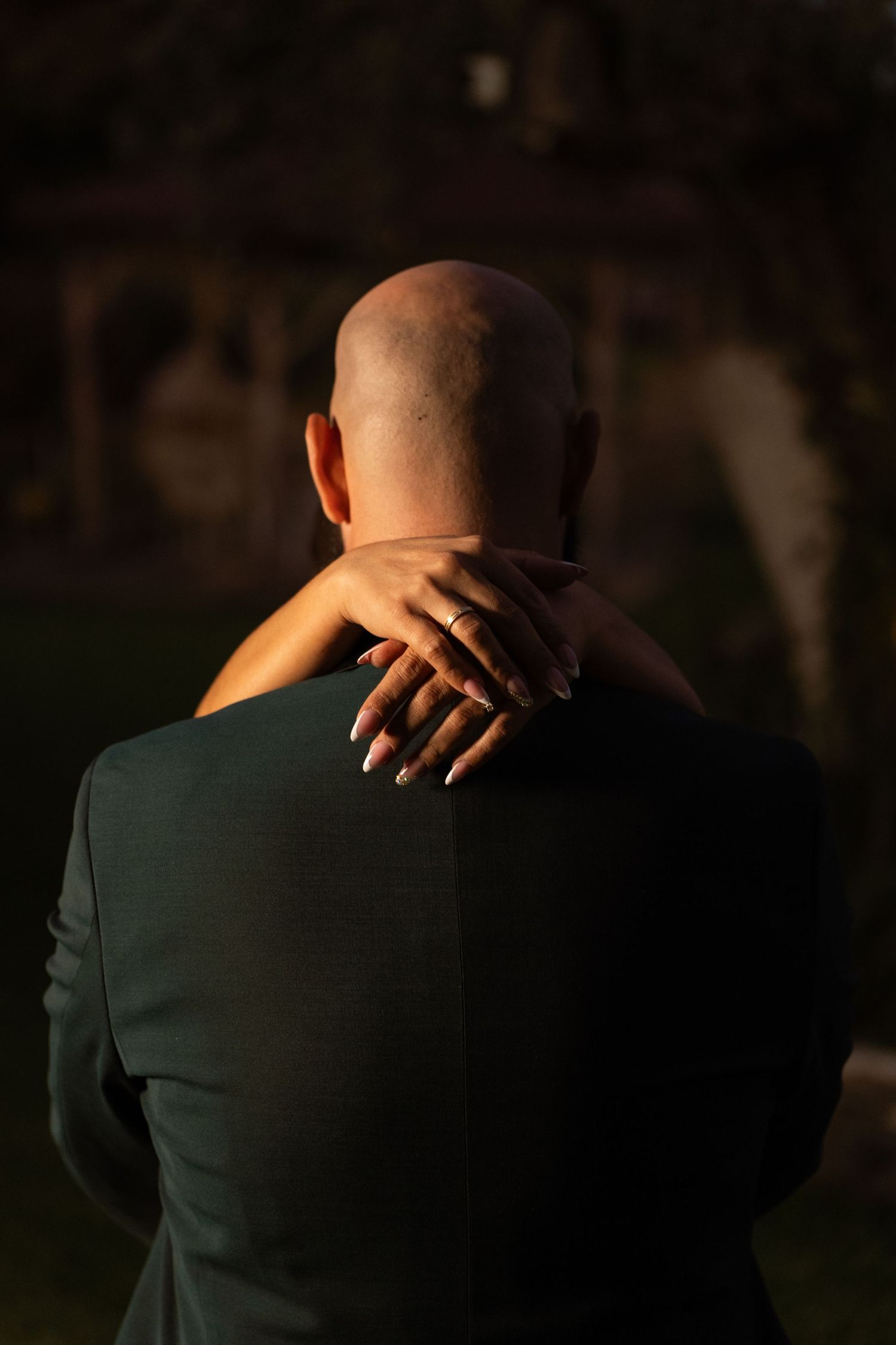 Back view of someone in a dark suit with hands clasped behind their bald head in warm lighting.