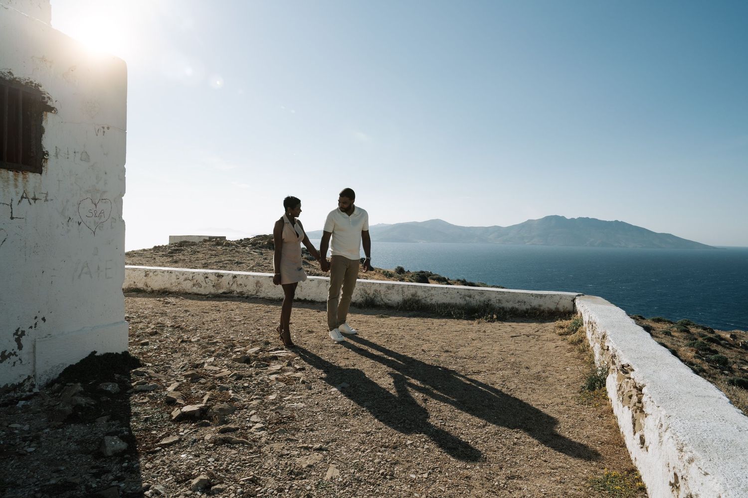 Couple walking along a white wall with mountains and ocean in the distance in Mykonos island