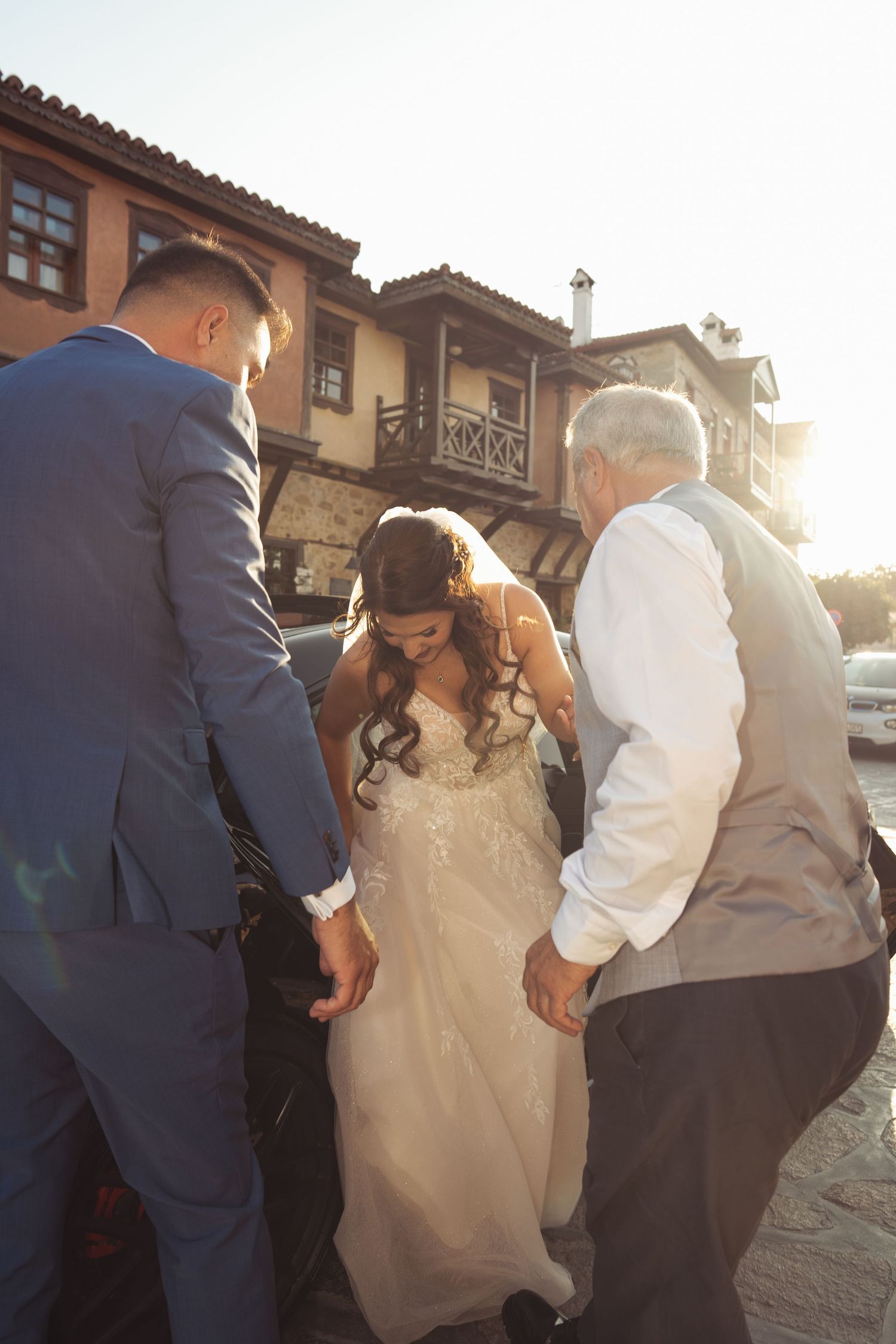A bride in a white wedding dress gets help with her dress train on a stone patio at sunset.