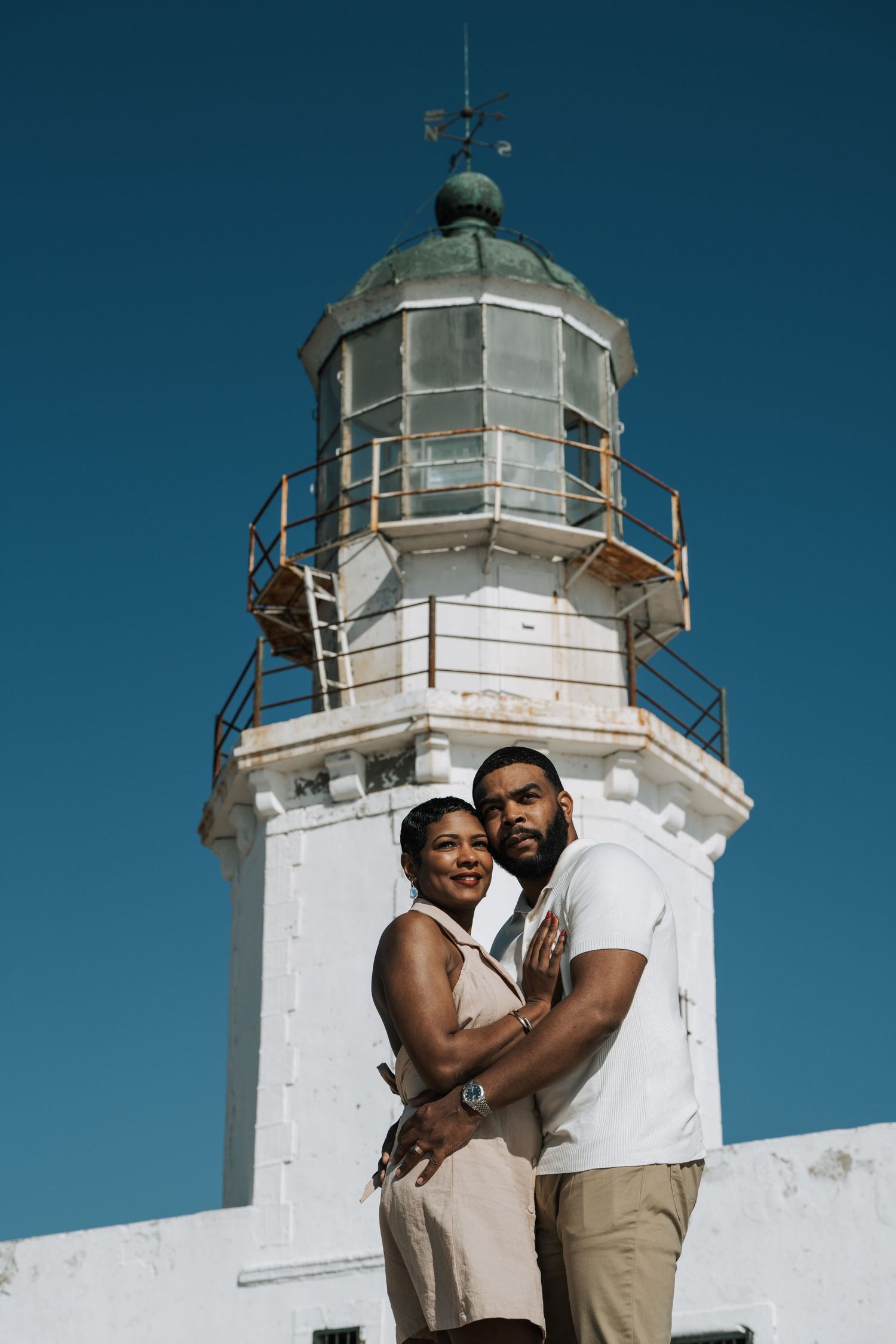 A romantic couple embraces in front of a white lighthouse against a deep blue sky in Mykonos Greece.