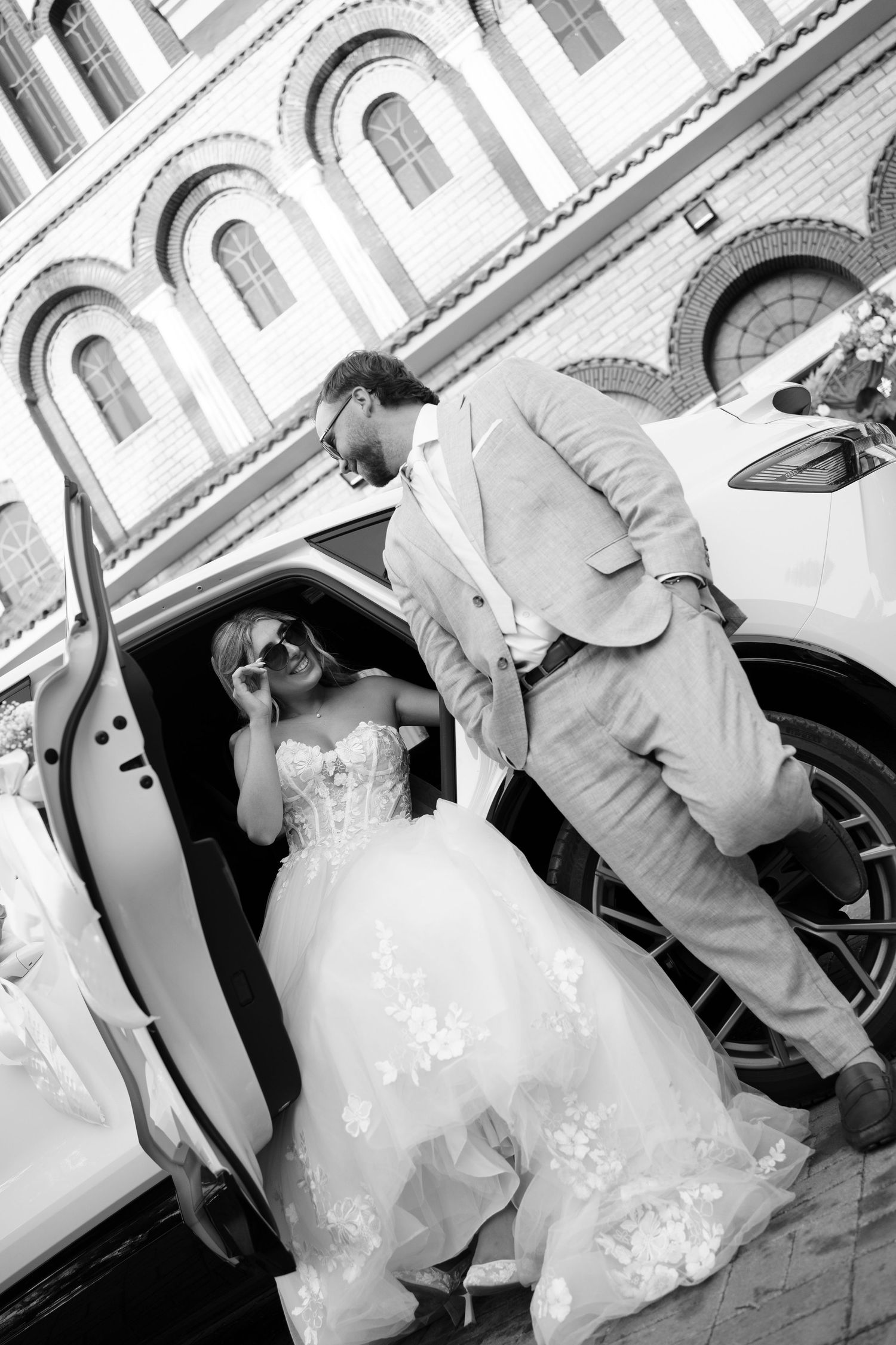 Wedding party assisting with dress while entering vehicle outside ornate building.