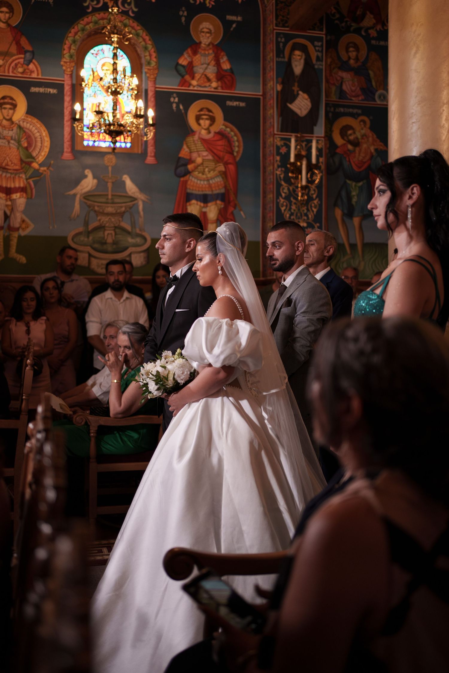 A wedding ceremony takes place in an ornate Orthodox church with religious icons and stained glass adorning the walls.