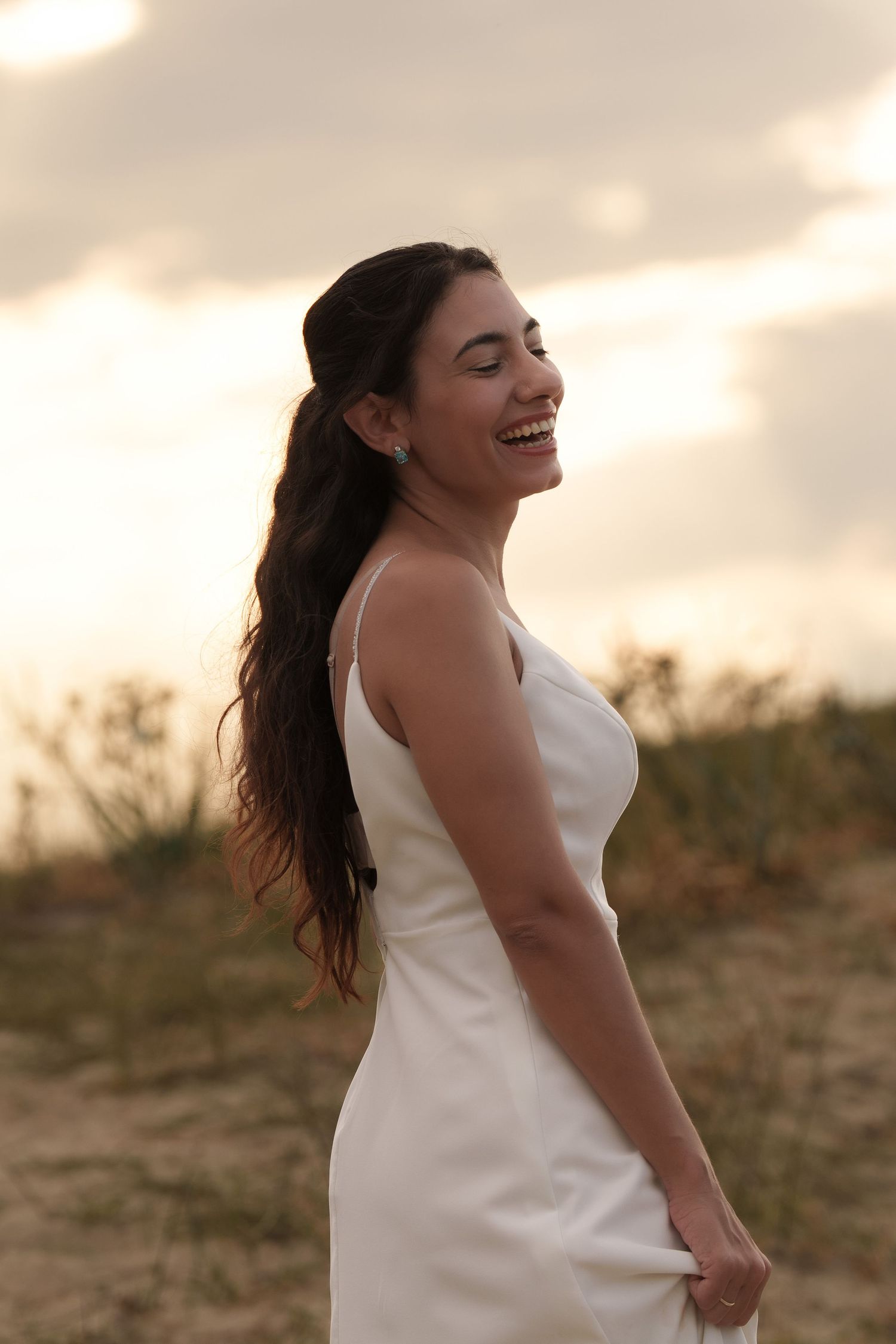 Woman in white sleeveless dress laughs joyfully during golden hour sunset photoshoot in outdoor setting.