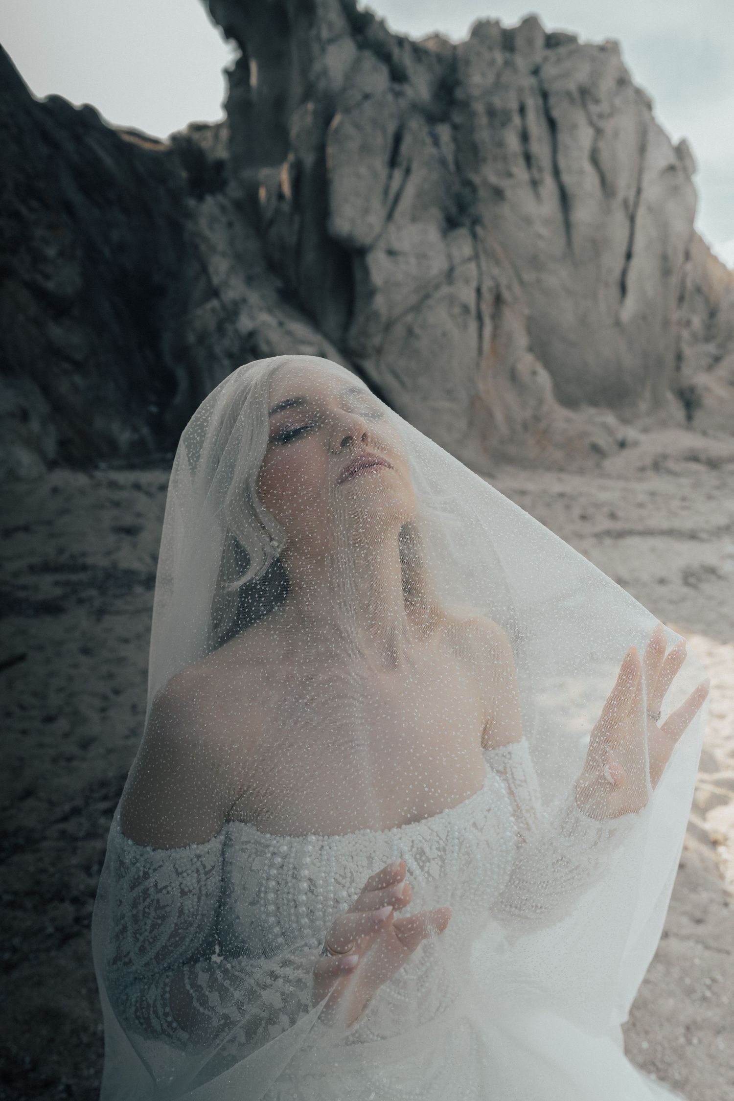 Artistic bridal portrait with veil against dramatic rocky landscape.