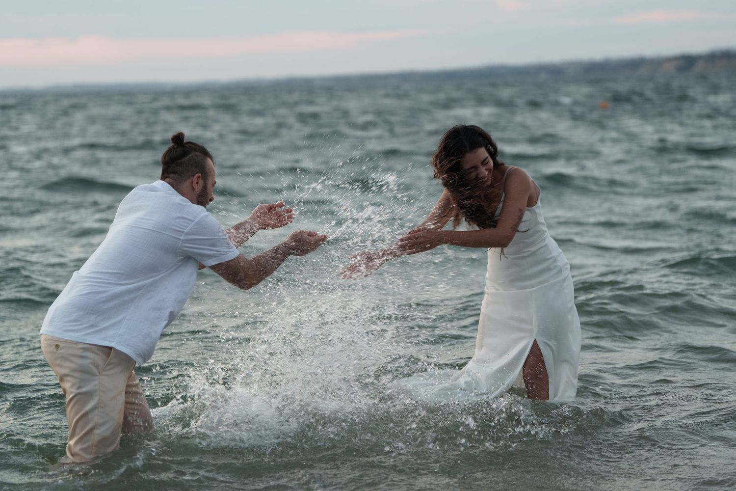 A couple in white and beige attire playfully splashing water at each other during a beach photoshoot.