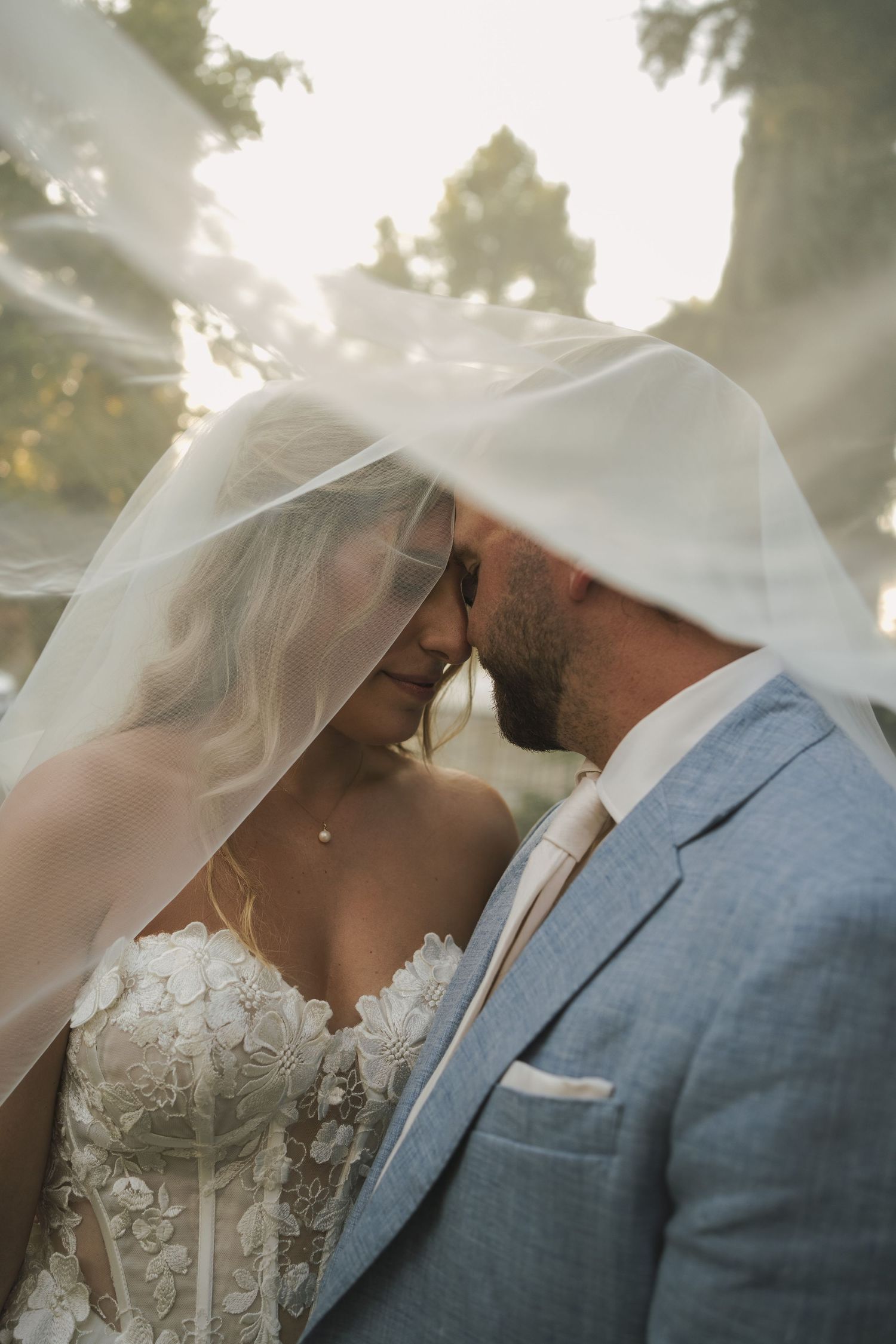 A tender moment shared under a flowing veil with sunlight streaming through trees.