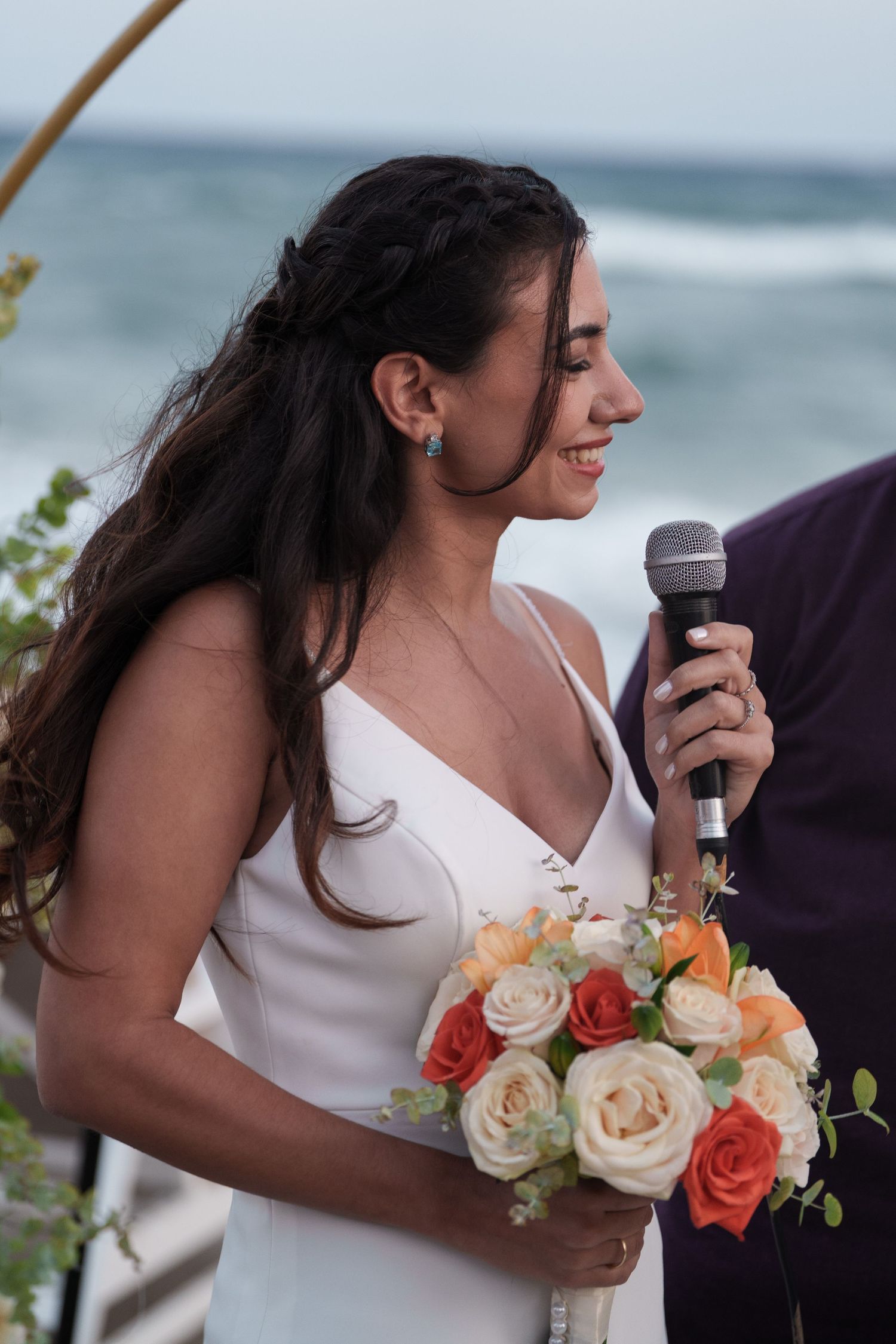 A bride in a white dress holds a microphone and a peach and coral rose bouquet at a beachside wedding ceremony.