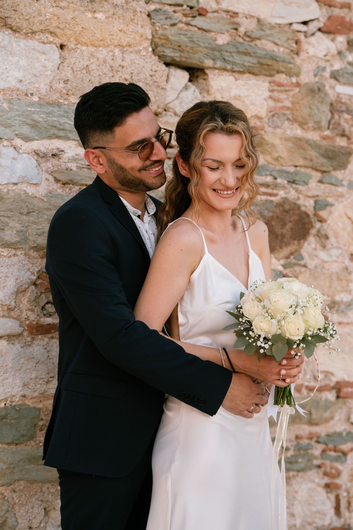 Romantic wedding couple embraces against rustic stone wall with white bouquet.