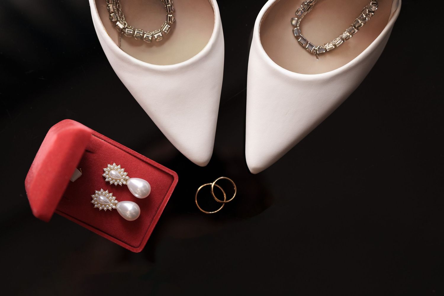 Wedding accessories including white shoes, rings and jewelry on dark background.