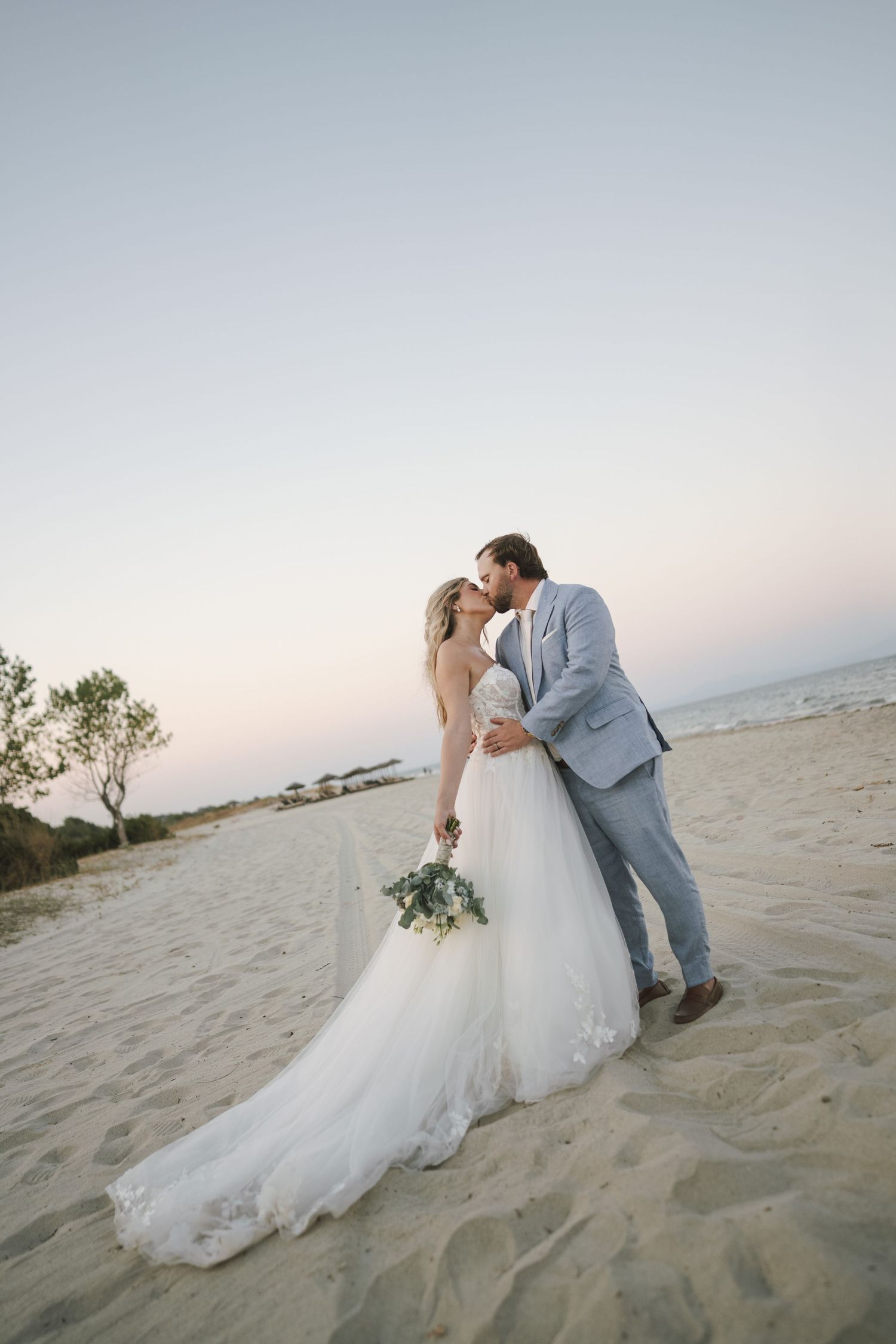 Romantic beach wedding portrait of couple kissing on sandy shore at sunset.