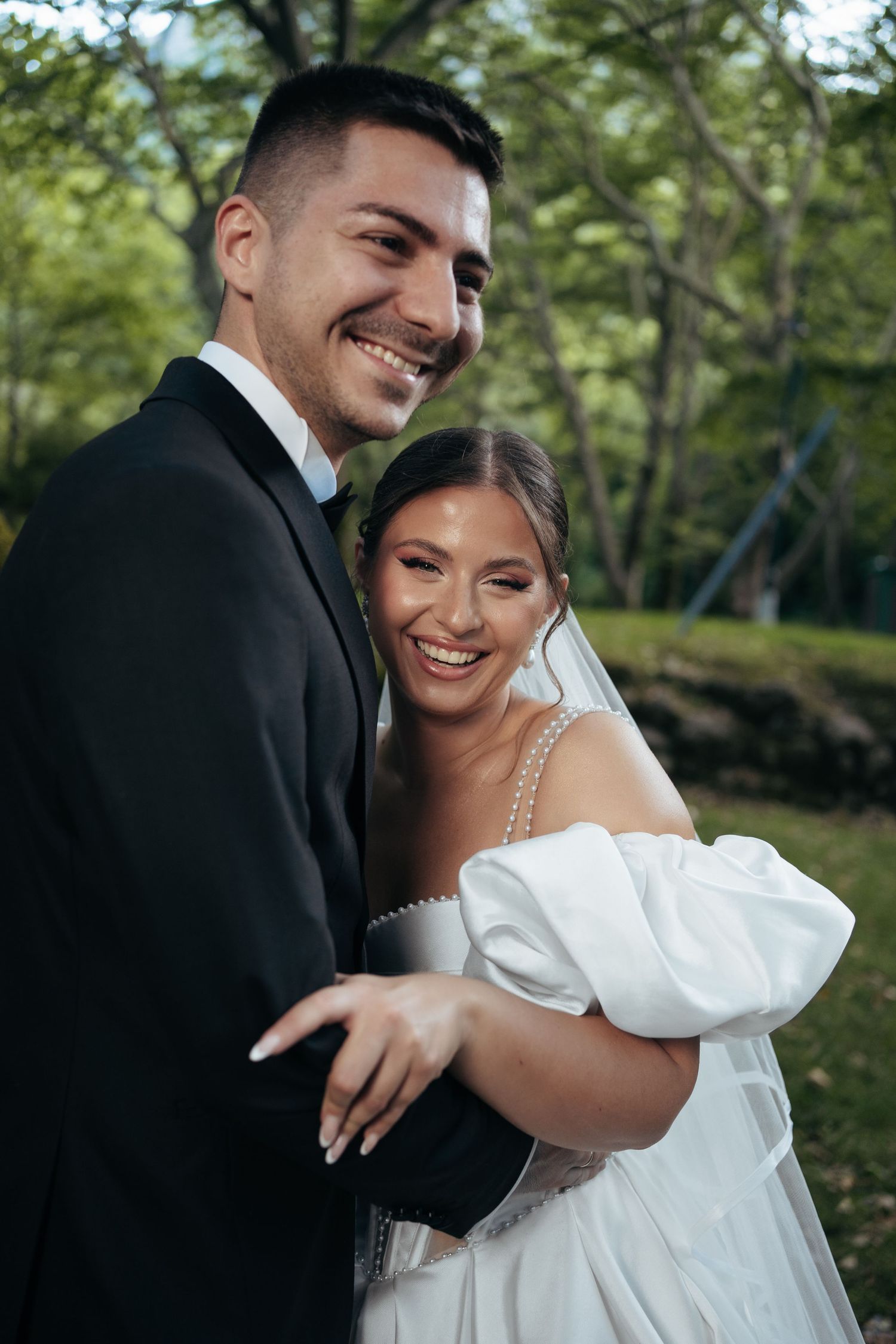 Joyful wedding couple embraces outdoors in a green garden setting wearing traditional formal attire.