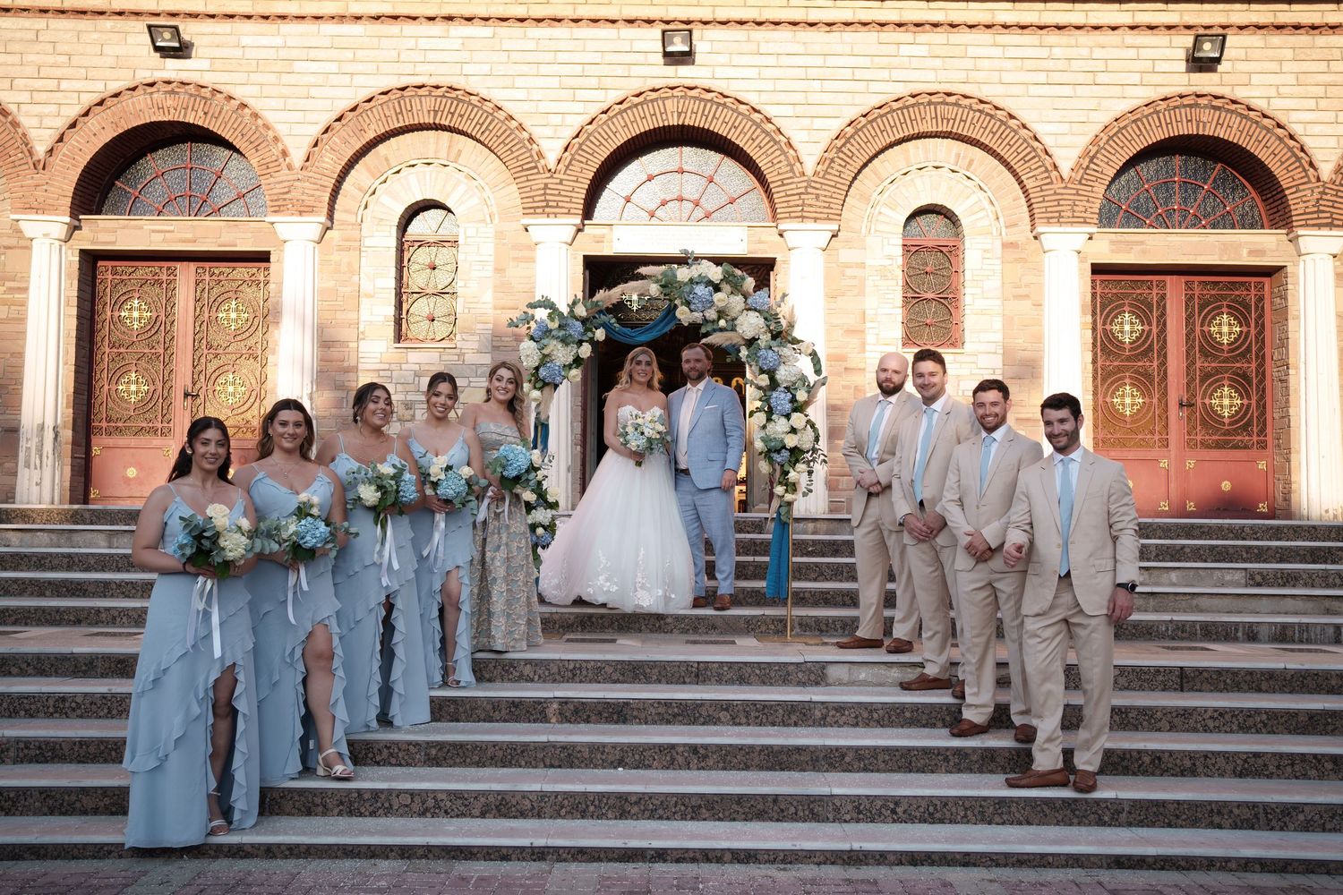 A wedding party poses on ornate stone steps outside a historic church building with arched doorways and decorative architecture.