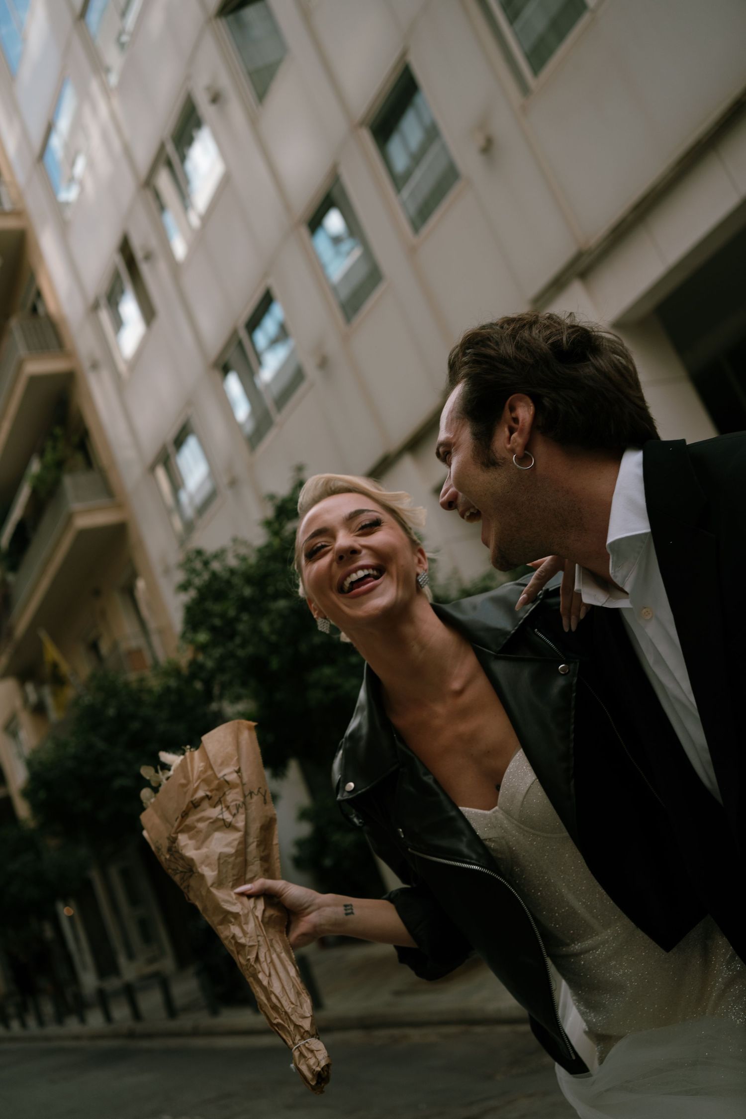 A joyful couple shares a laugh while holding a bouquet on an urban street in Athens Greece