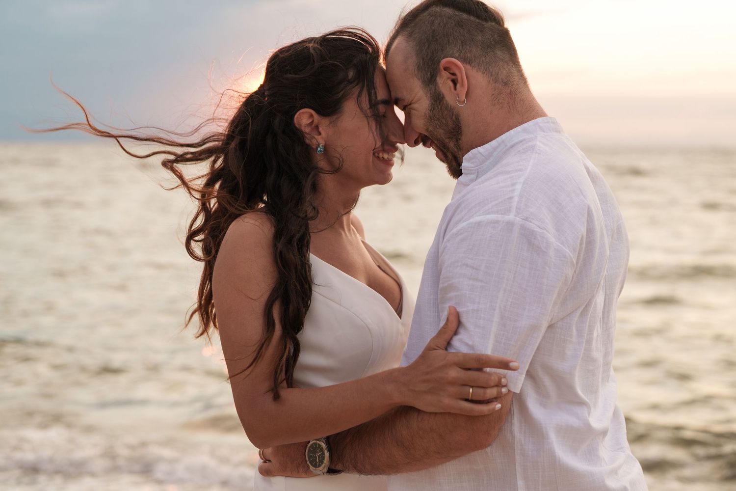 A romantic couple embraces on the beach at sunset with waves in the background.