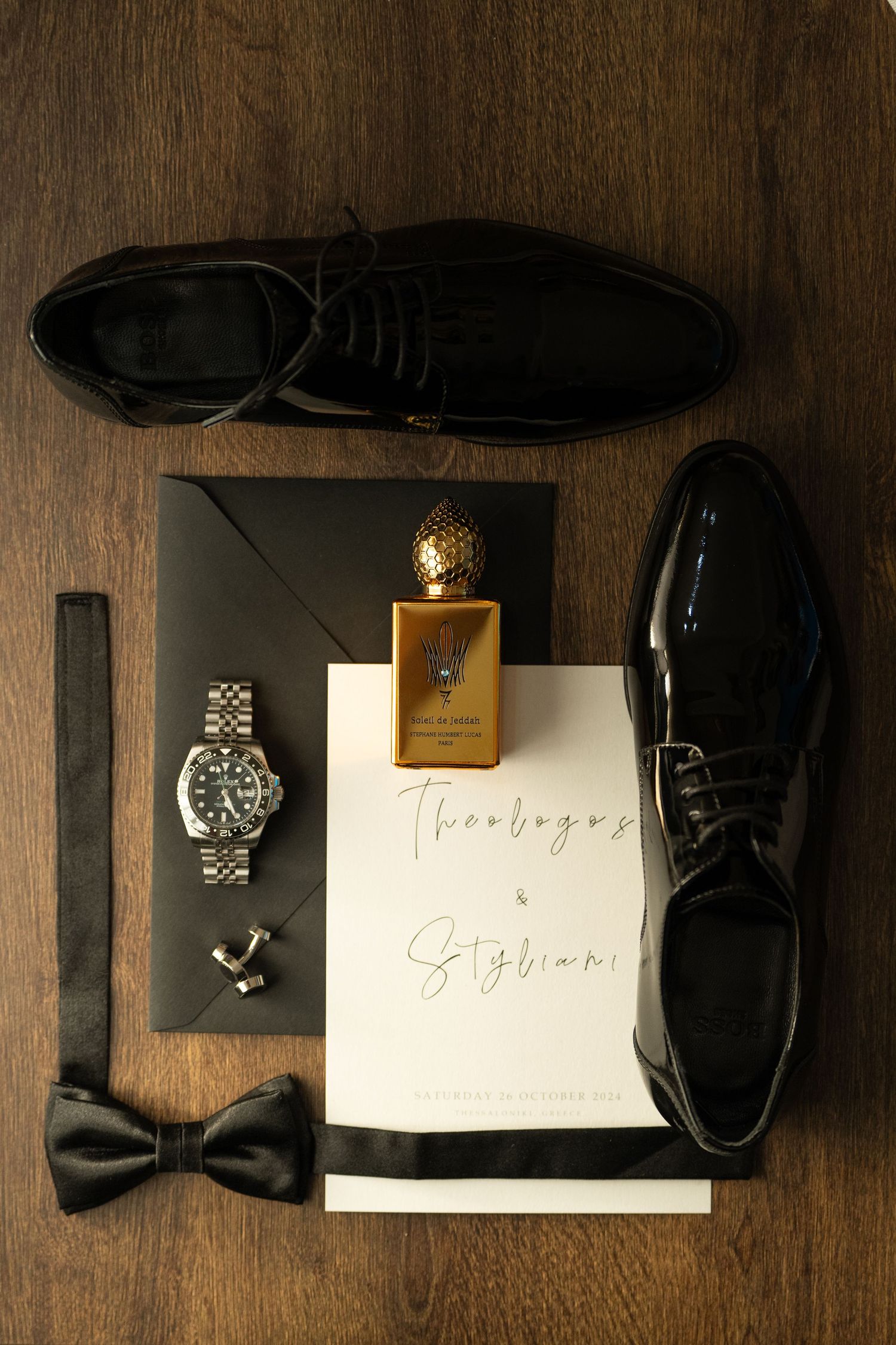 Flat lay of groom's accessories including shoes, bow tie, watch and wedding stationery on wooden surface.