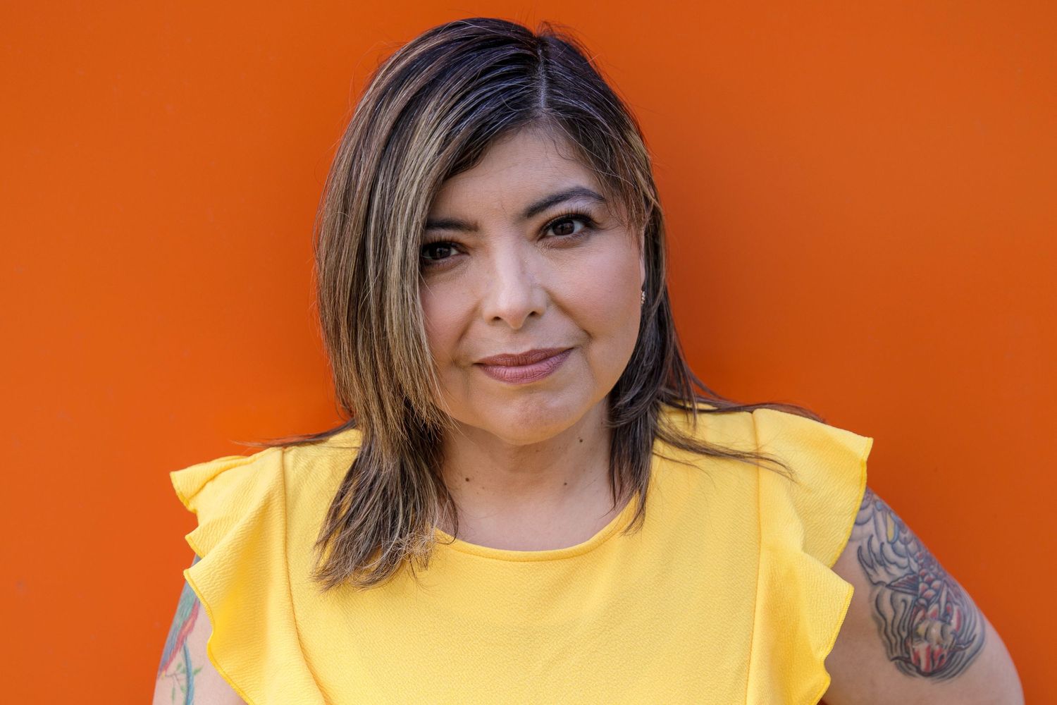 Portrait of person in yellow ruffled top with tattoo against orange backdrop.