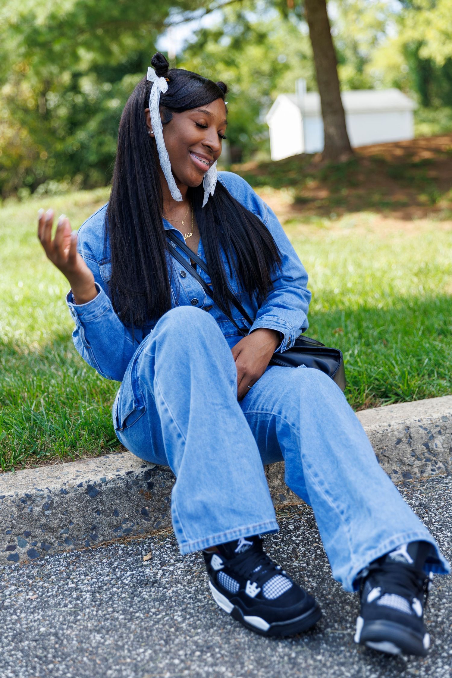 A casual outdoor portrait showing someone in a denim outfit sitting on a curb in a park setting.