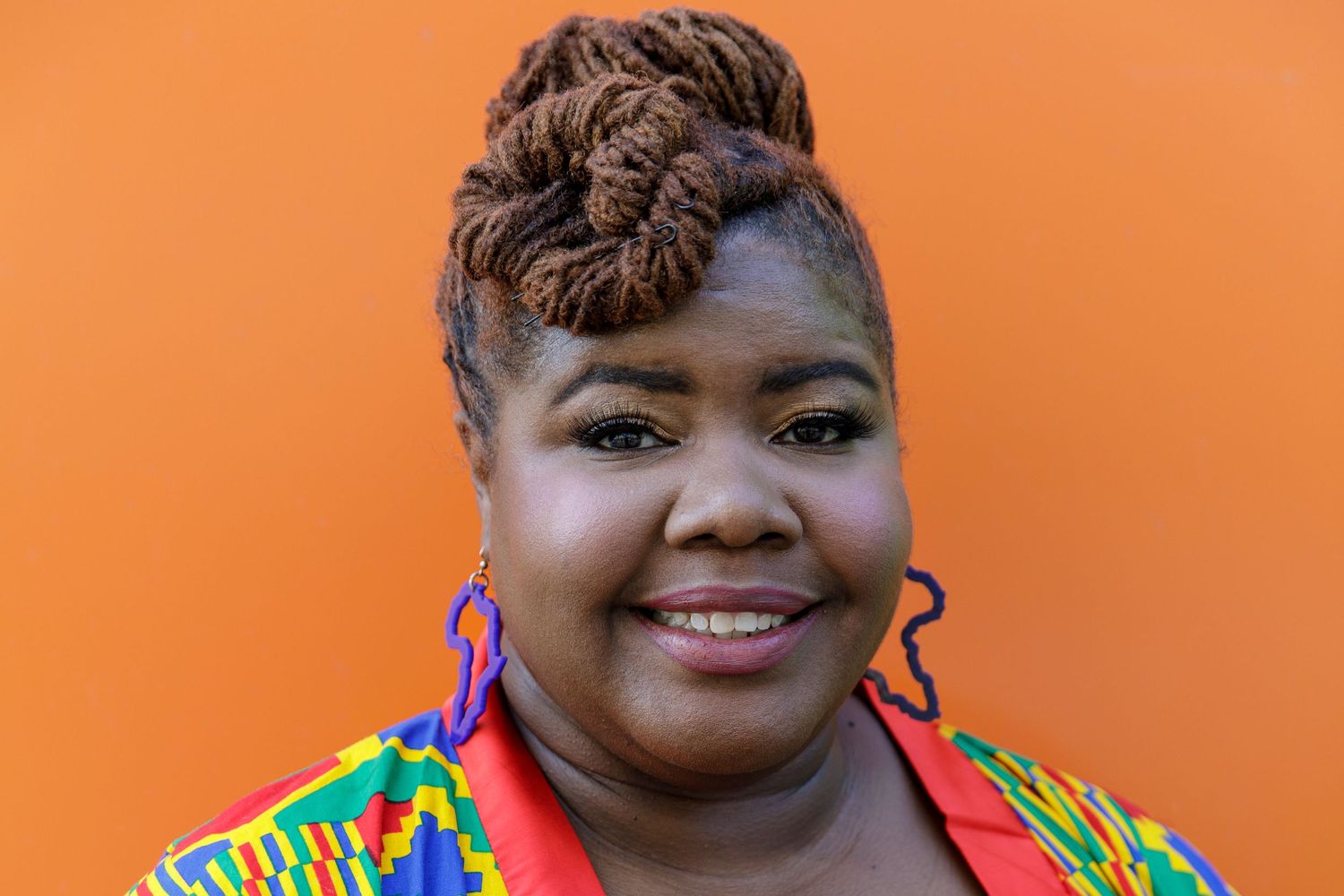 Portrait of person in colorful patterned top with styled hair against orange background.
