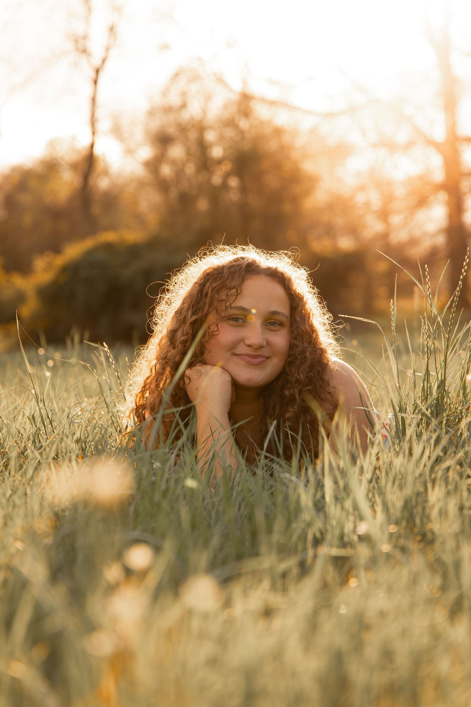 Young woman with curly hair lying in golden grass field during sunset, smiling warmly at camera.