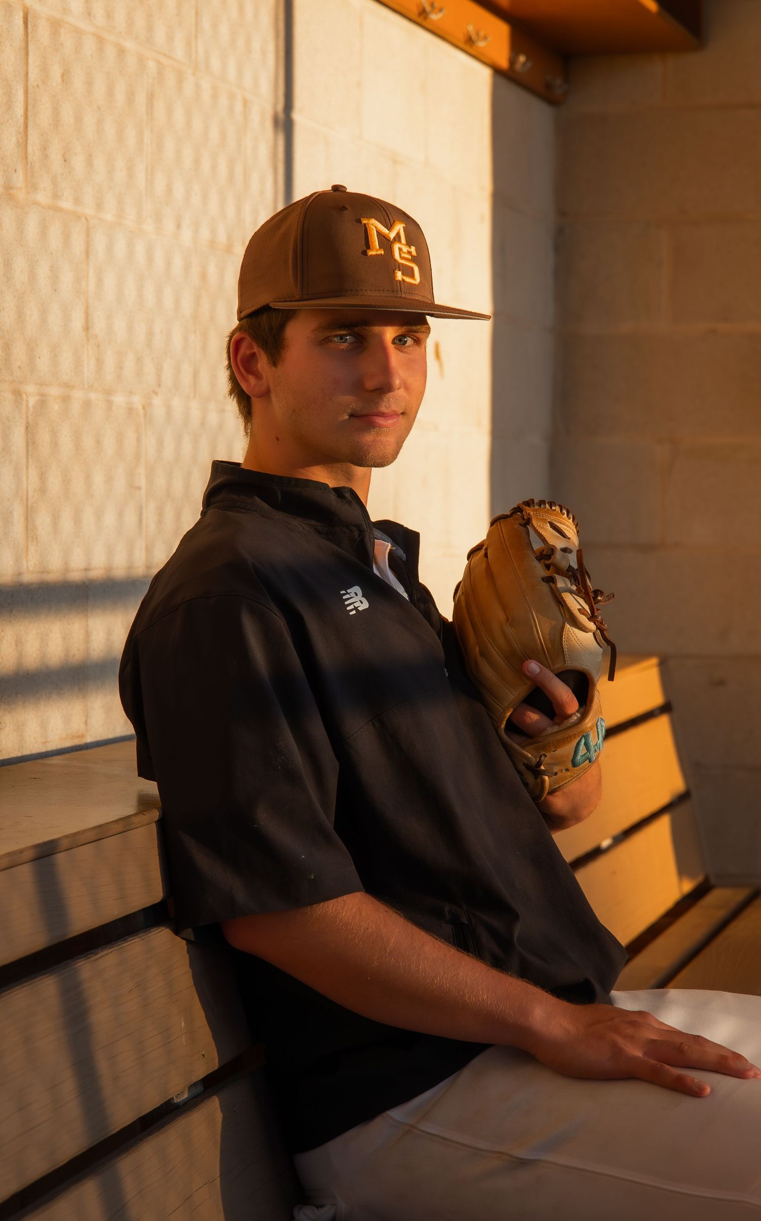 Young baseball player in brown MS cap and black New Balance shirt holds glove in dugout.