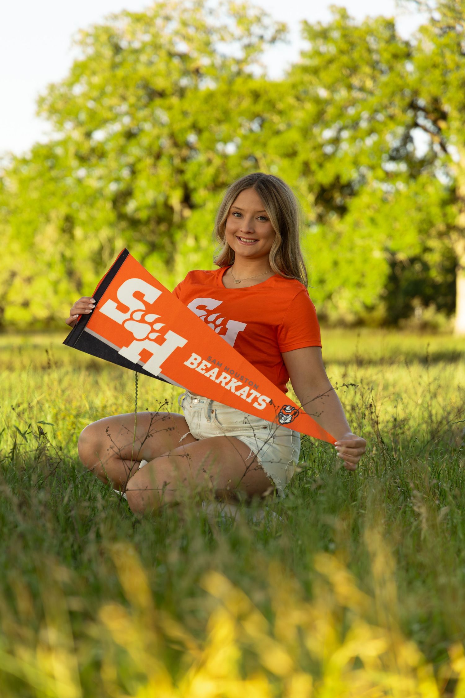 Young woman in orange shirt holding Sam Houston State Bearkats pennant, sitting in green grass field.