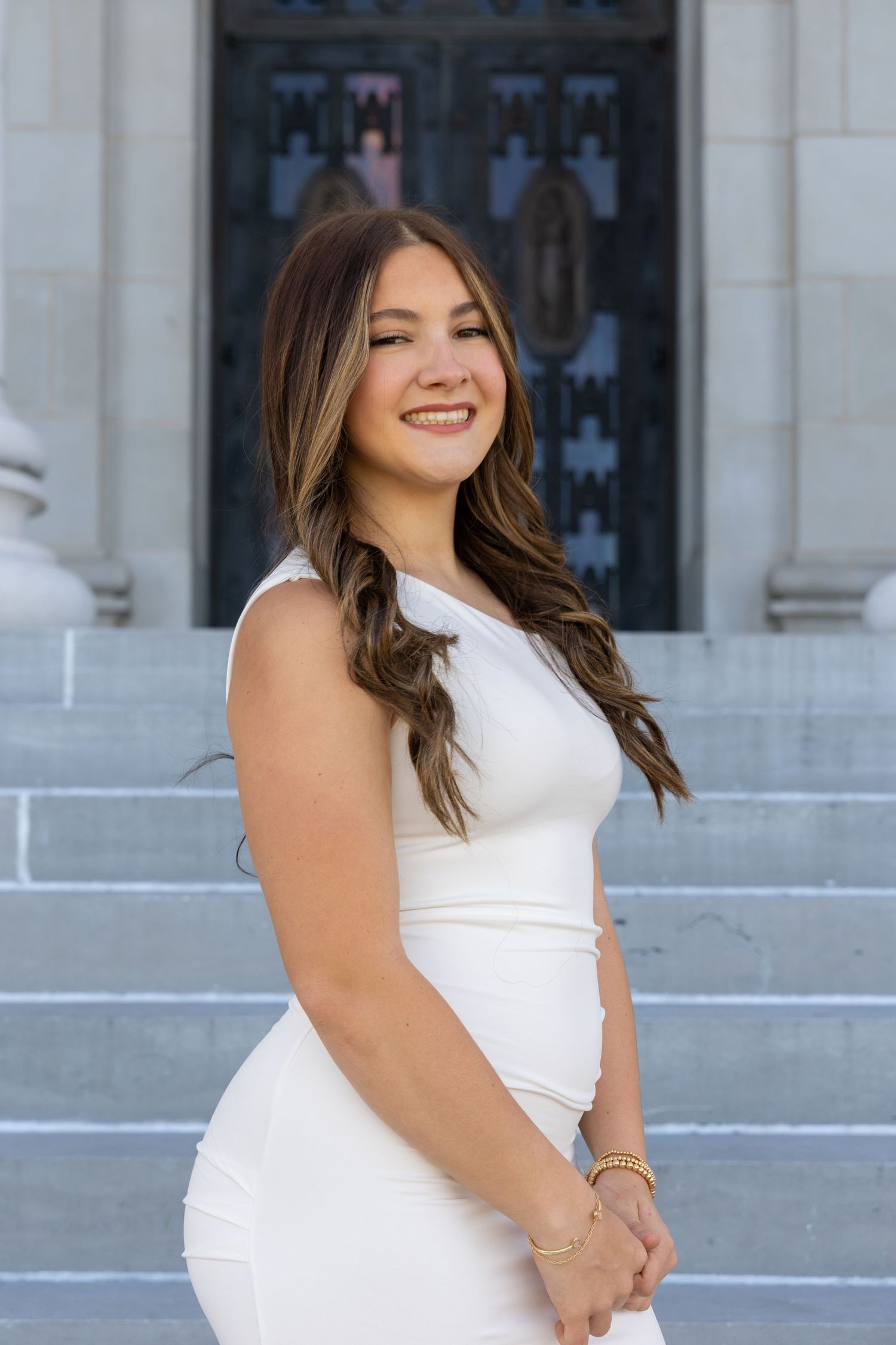 Portrait of someone in a sleeveless white form-fitting dress standing on stone steps with a gothic door in background.