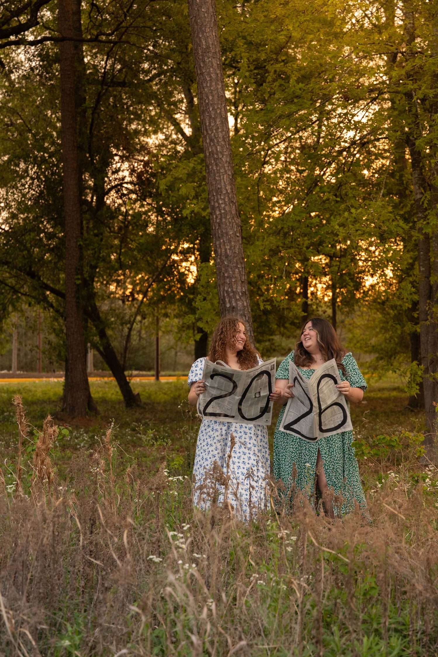 Two young women in floral dresses hold 2026 signs in a sunlit forest during golden hour.