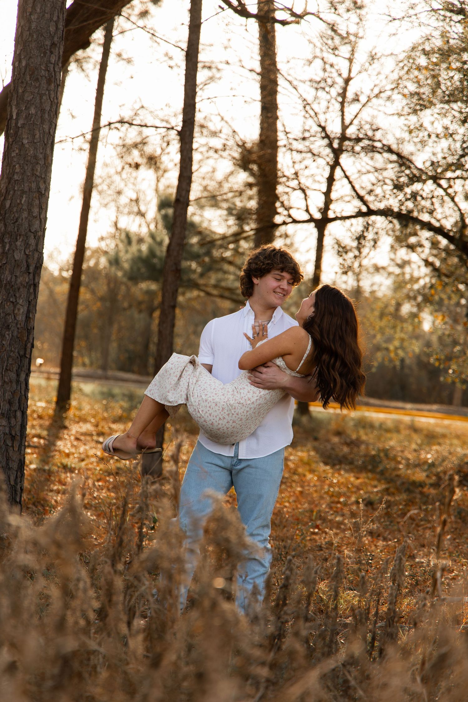 Young couple sharing a romantic moment in a sunlit autumn forest, man lifting woman in white dress among golden trees.