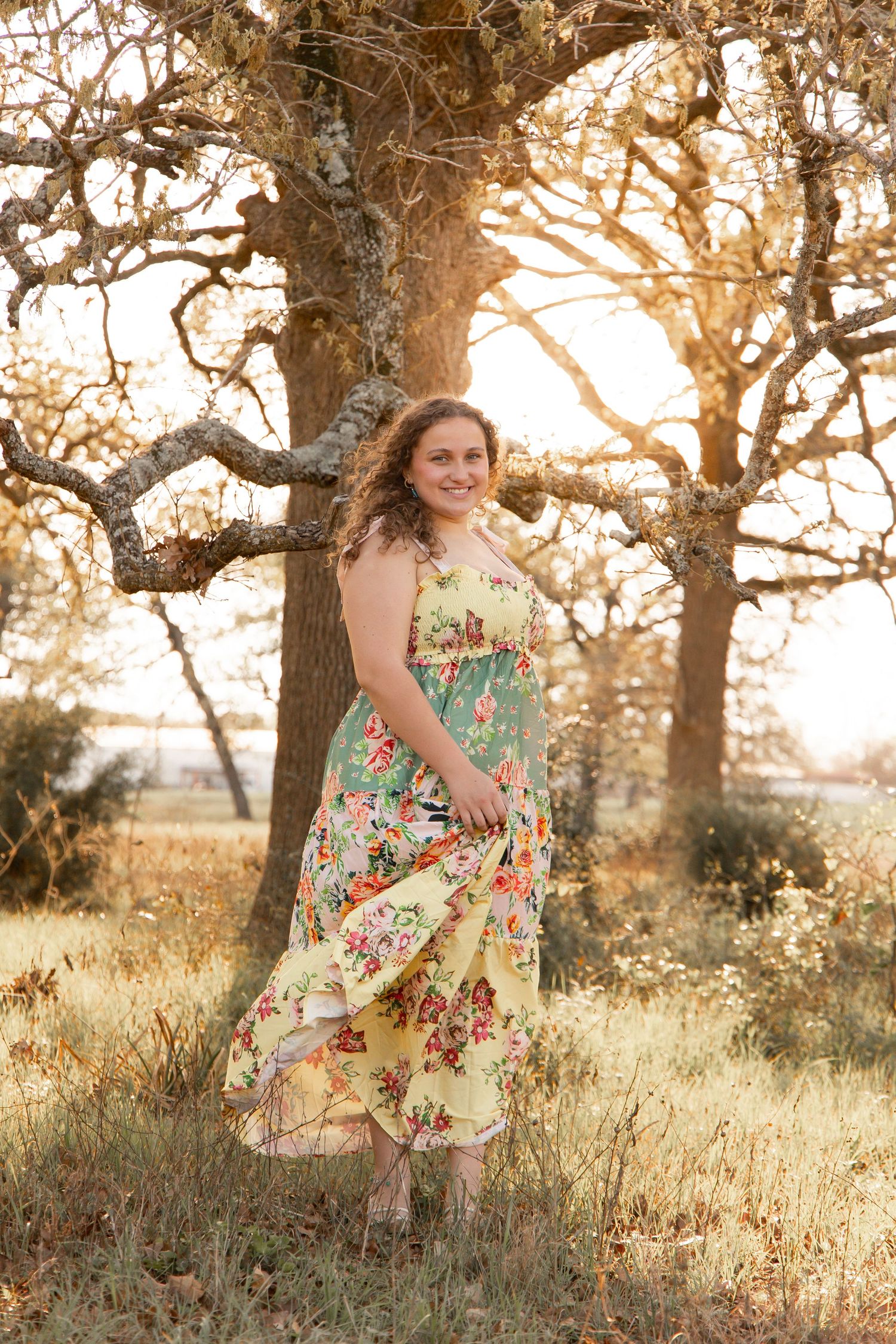 Young woman in floral yellow maxi dress poses outdoors near trees with golden sunset backlight.