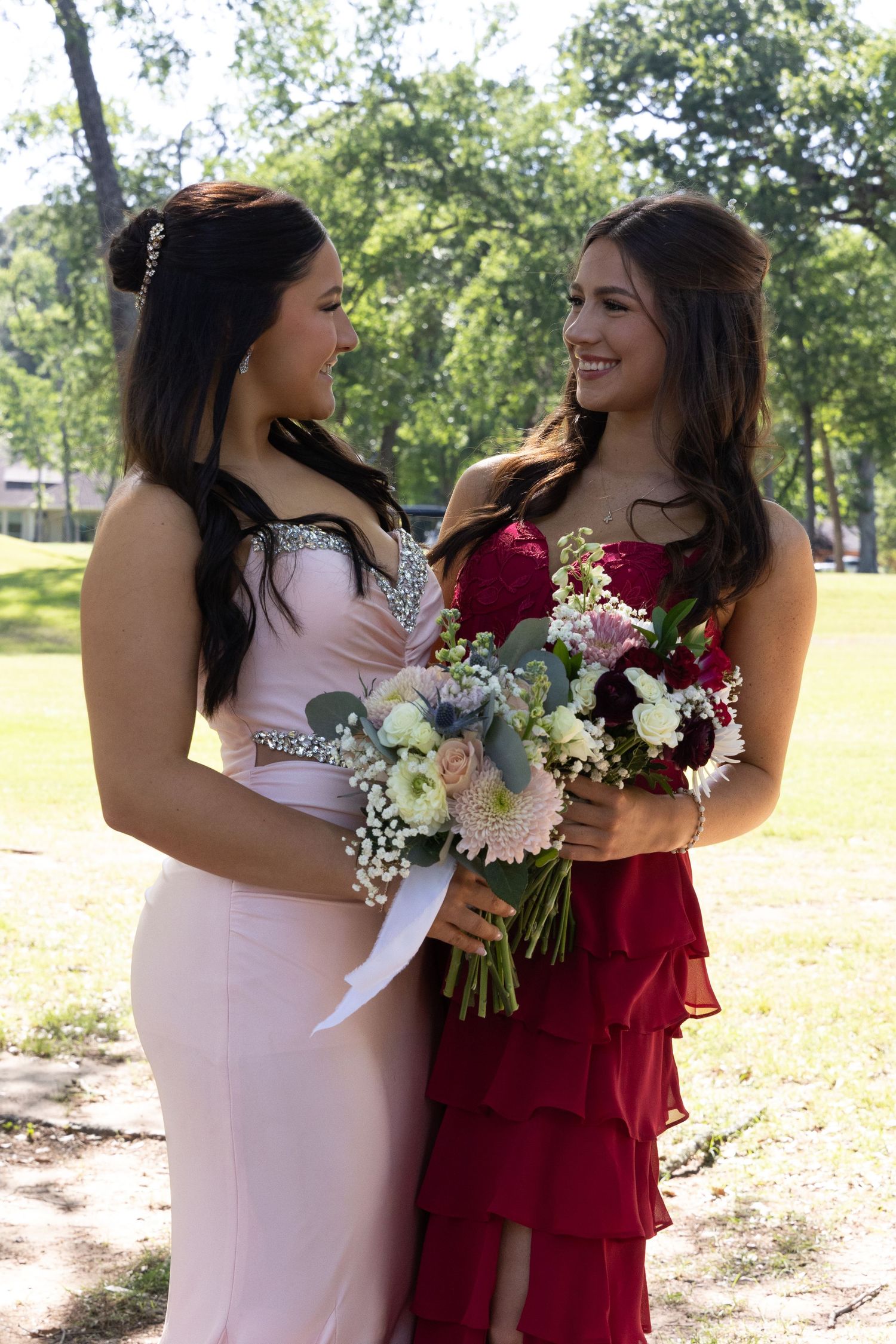 Two young women in elegant prom dresses holding bouquets, smiling at each other outdoors in a park.