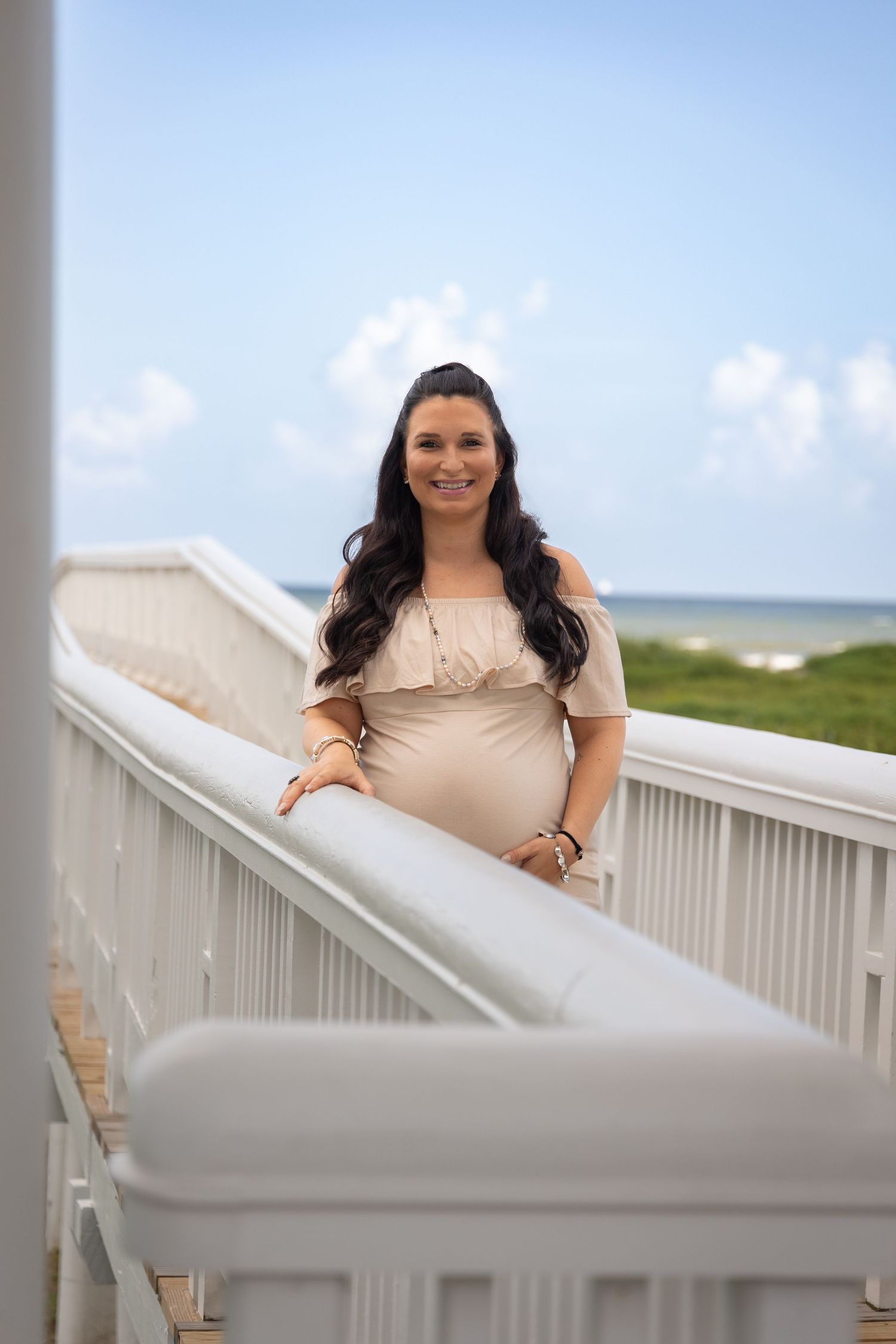 Smiling pregnant woman in beige off-shoulder dress poses on white beach boardwalk railing with ocean view behind her.