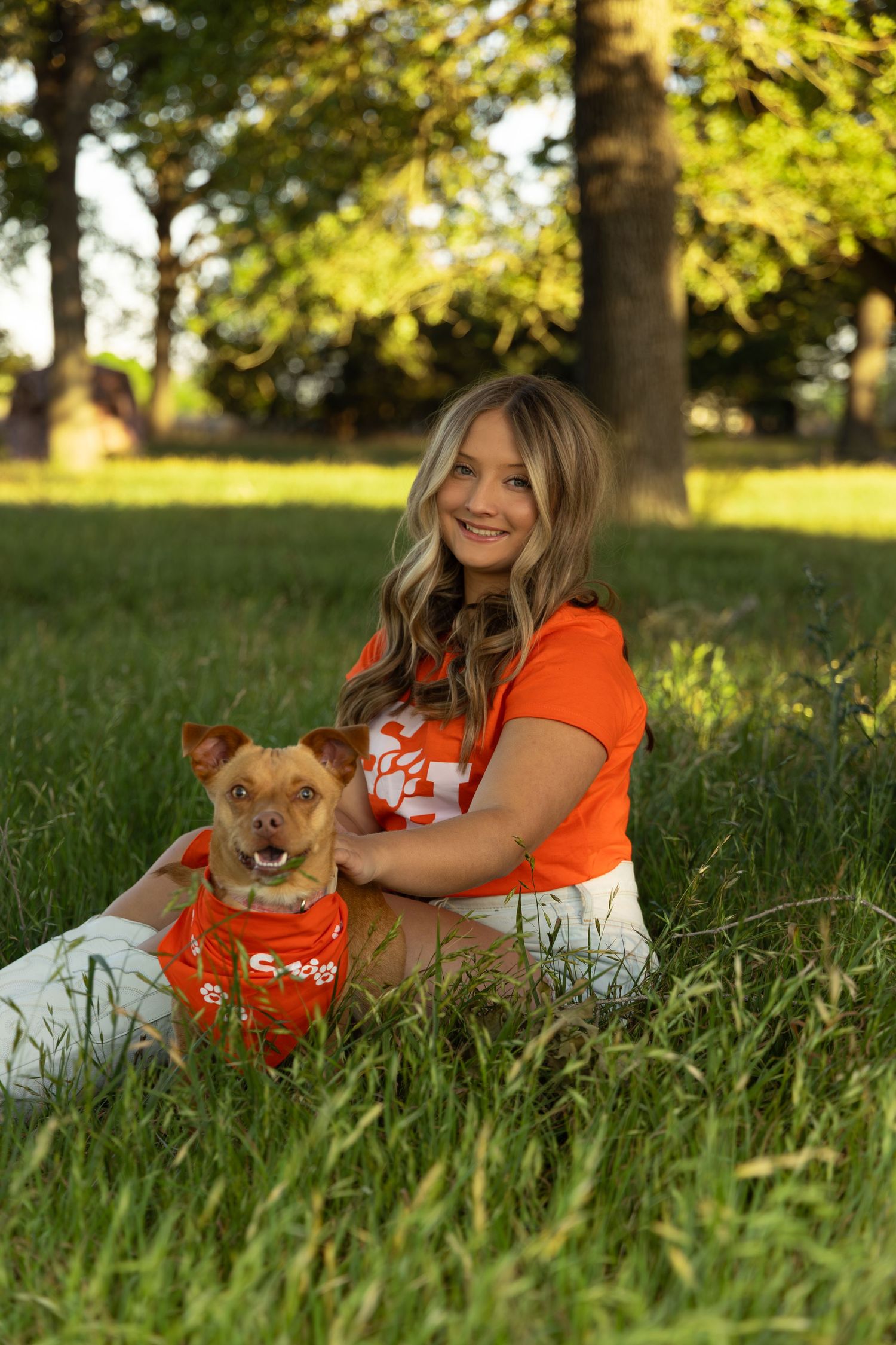 Smiling young woman in orange shirt sitting in grass with small brown dog wearing matching orange bandana in sunny park.