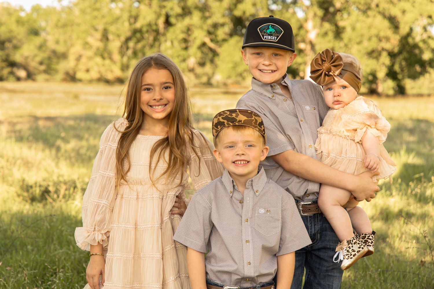 Four smiling children pose together in a sunny green meadow, dressed in coordinating neutral outfits for a family portrait.