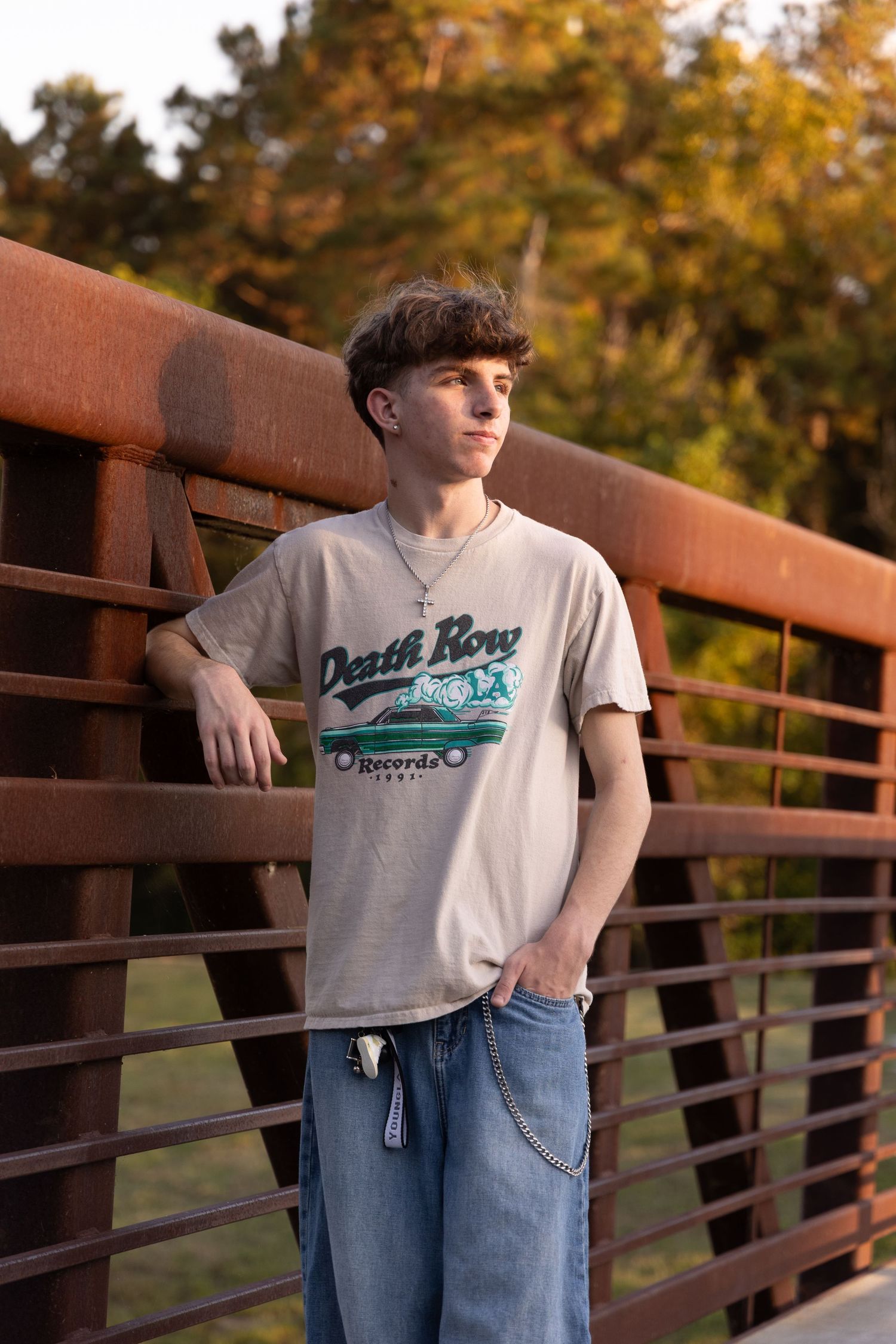 Teen boy in Death Row Records tee and jeans leaning on rustic bridge railing outdoors at golden hour.
