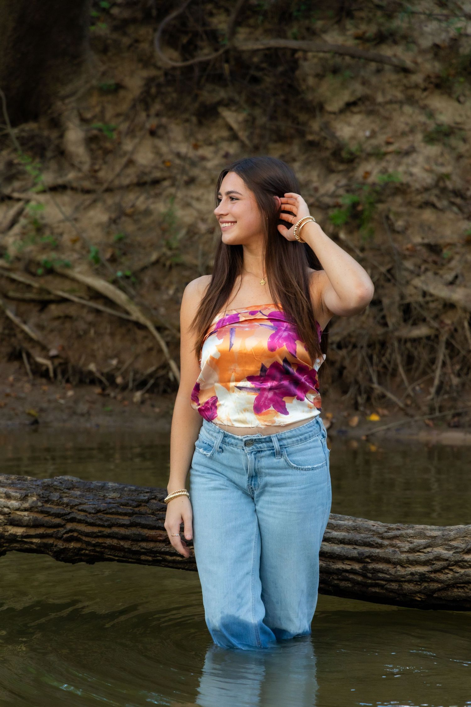 Young woman in floral tube top and jeans standing in a shallow creek near a fallen log in a natural setting.