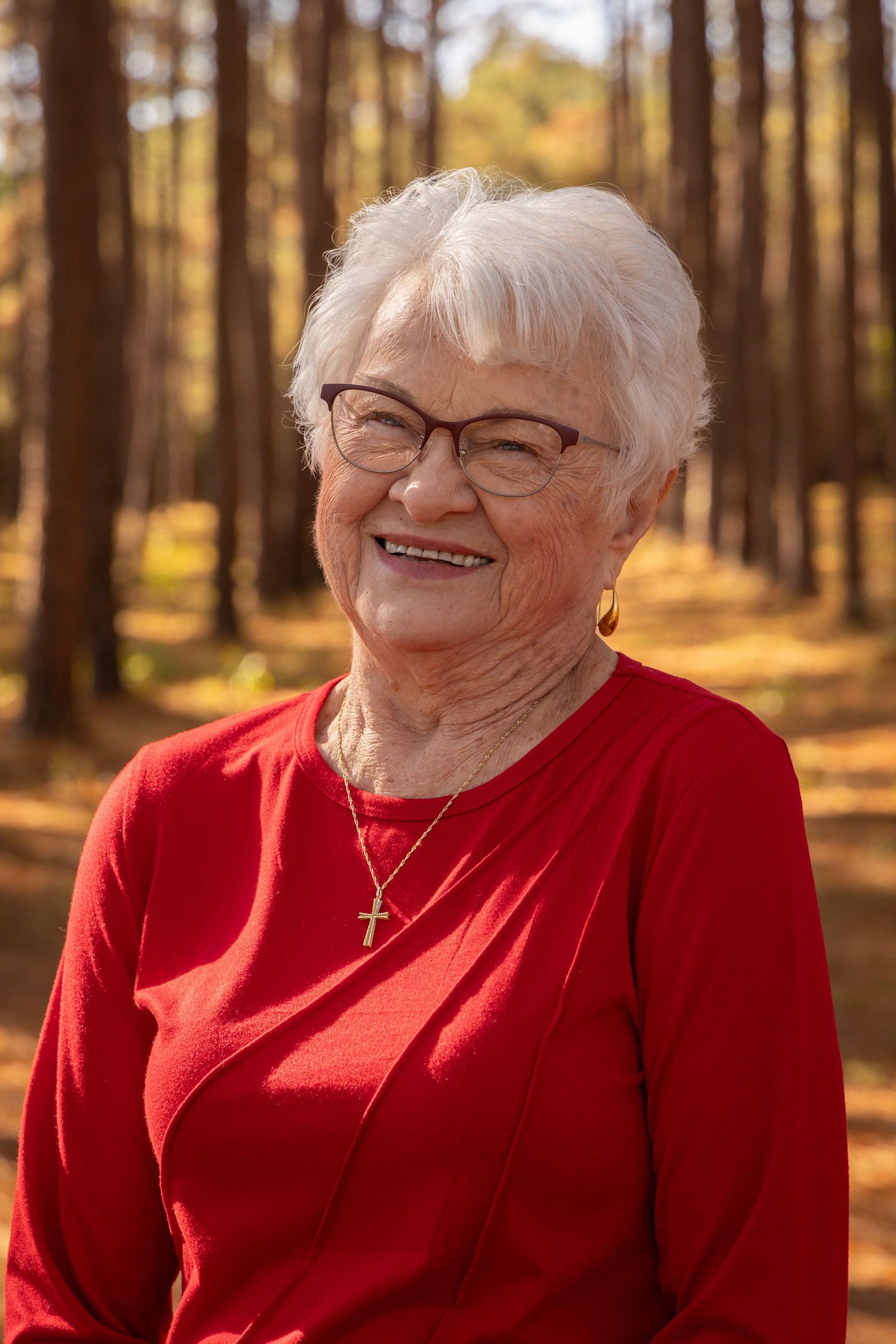 Smiling elderly woman with white hair and glasses wearing a red top and gold cross necklace outdoors in a sunny forest.