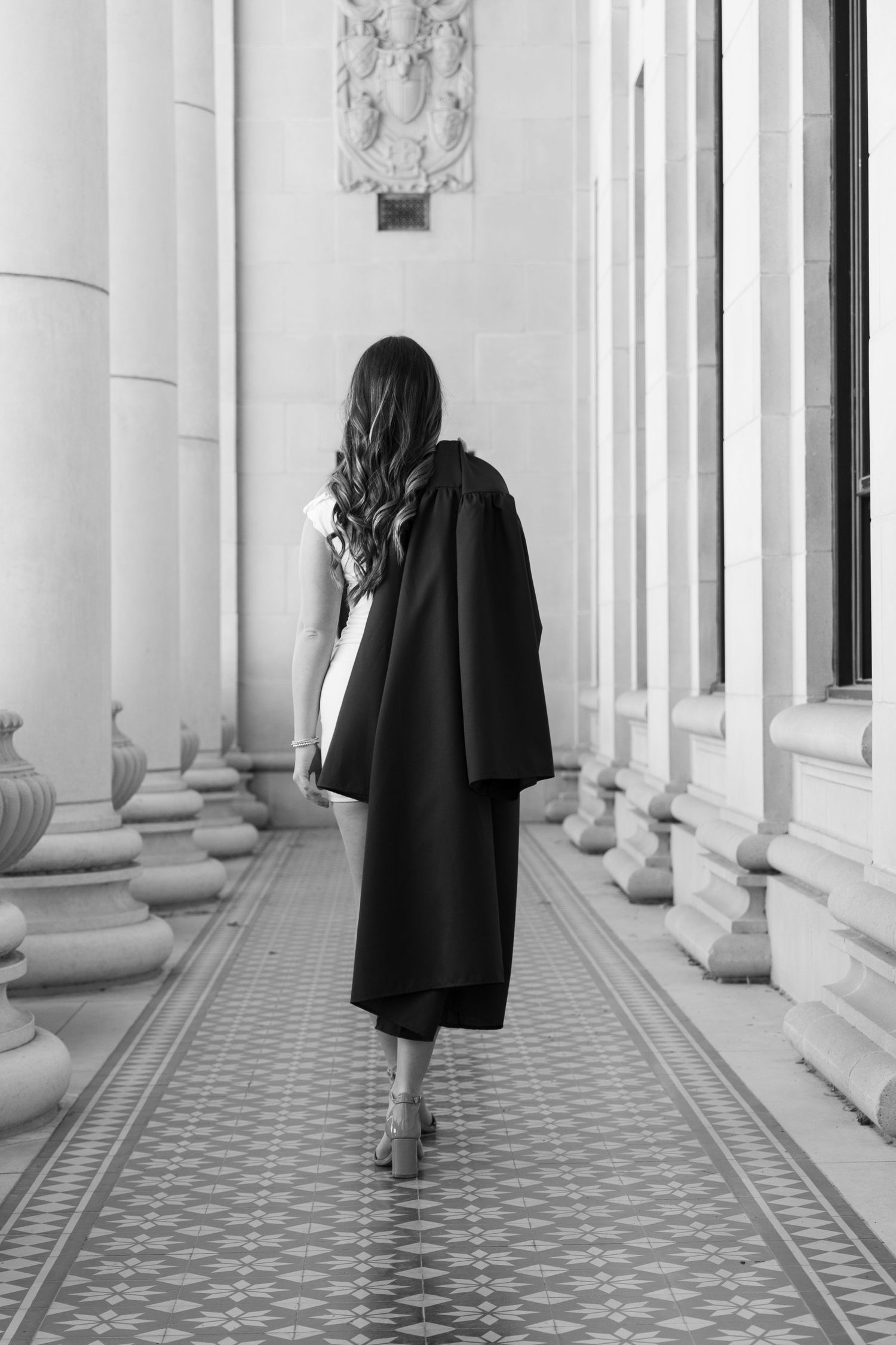 A black and white photo of someone in a long coat walking on a patterned floor along a classical colonnade hallway.