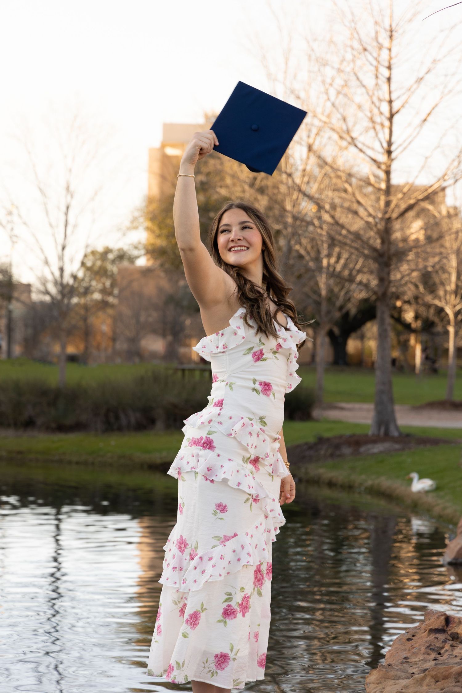 Young woman in floral dress celebrating graduation by holding diploma aloft near a pond in a park.