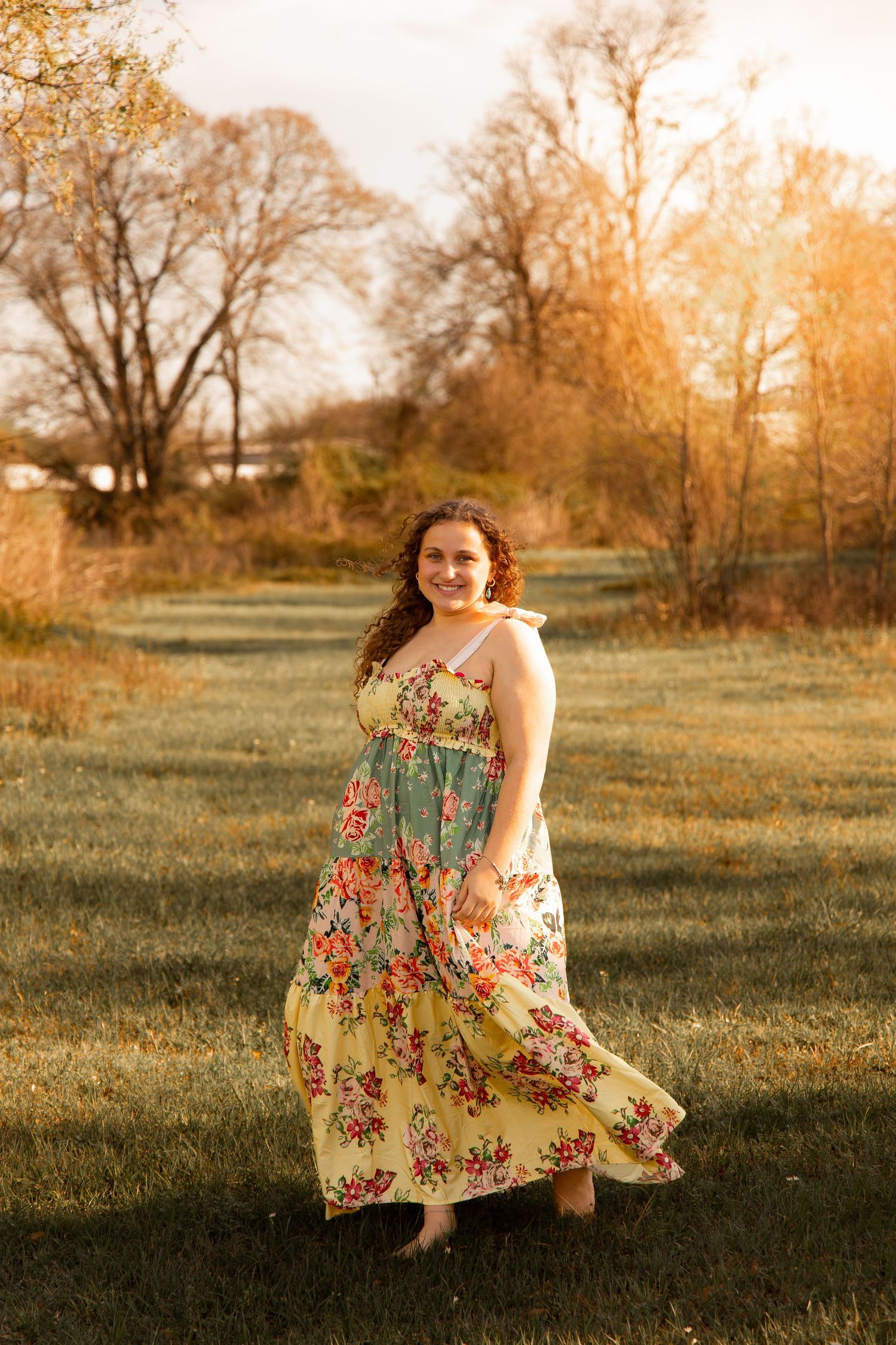 Young woman in floral maxi dress twirling in golden hour sunlight on green grass with autumn trees in background.
