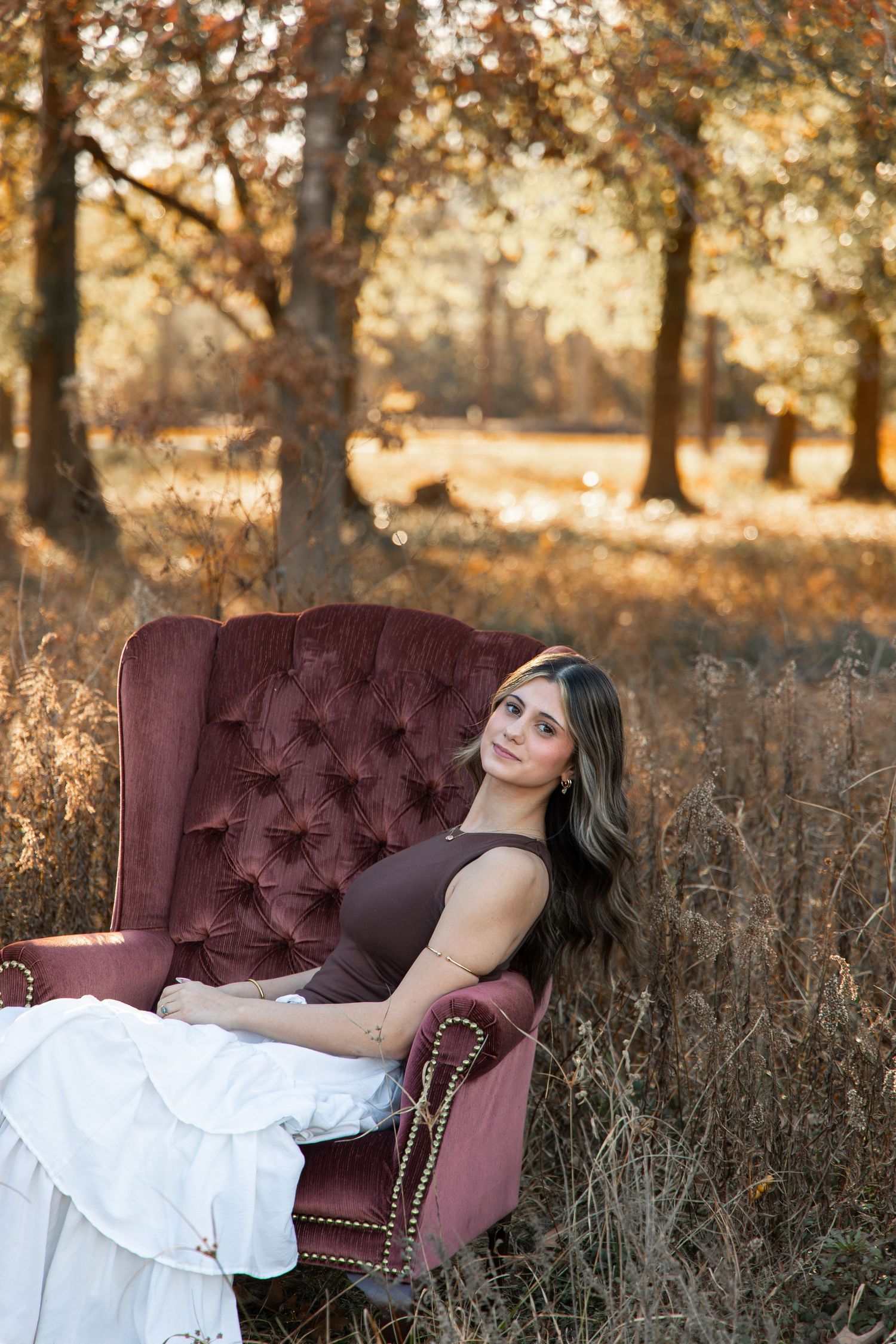 Woman in brown top and white skirt relaxing in a tufted velvet armchair outdoors in an autumn field.